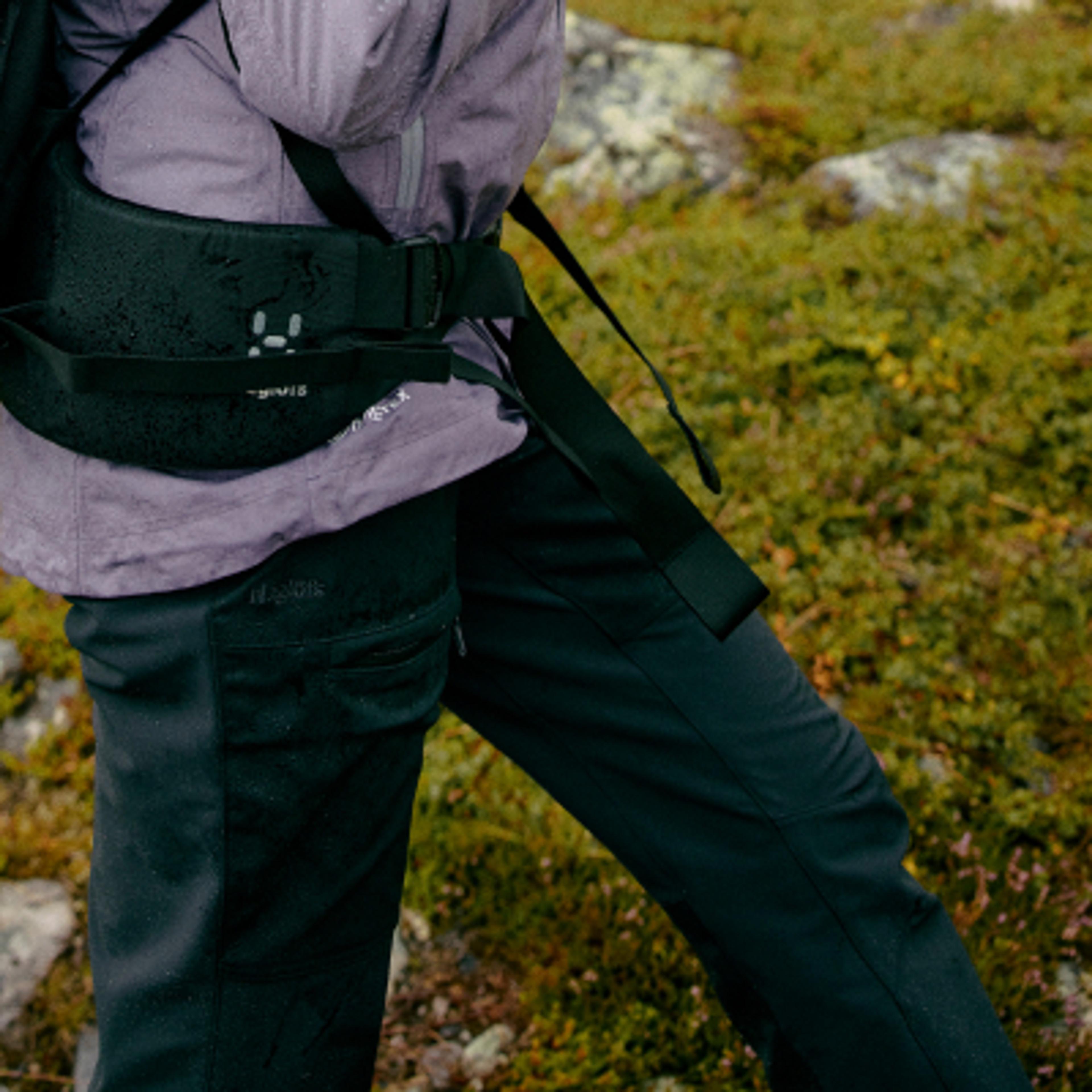 A person walks outdoors wearing a wet purple jacket, black Haglöfs waist pack, and black pants.