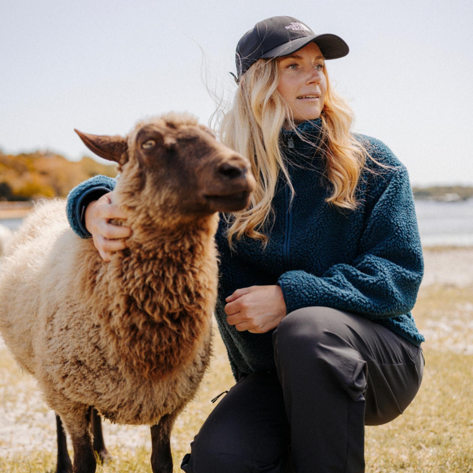 A woman in a teal fleece jacket kneels, petting a light brown sheep with a dark face outdoors.