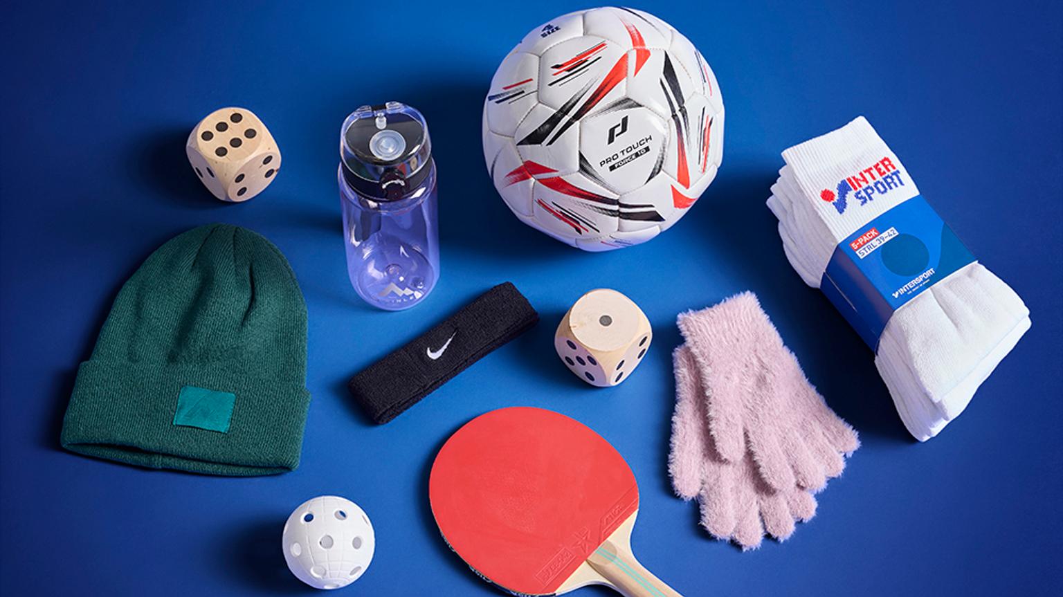 Various sports and casual items arranged on a blue background: a soccer ball, stacked white socks, a green beanie, pink gloves, a purple water bottle, a black Nike headband, a red table tennis paddle, a small white ball, and two wooden dice.
