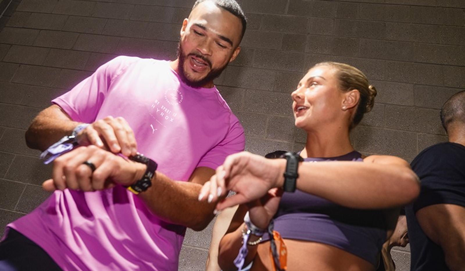 A man in a pink shirt and a woman in a purple sports bra look at their smartwatches and talk.