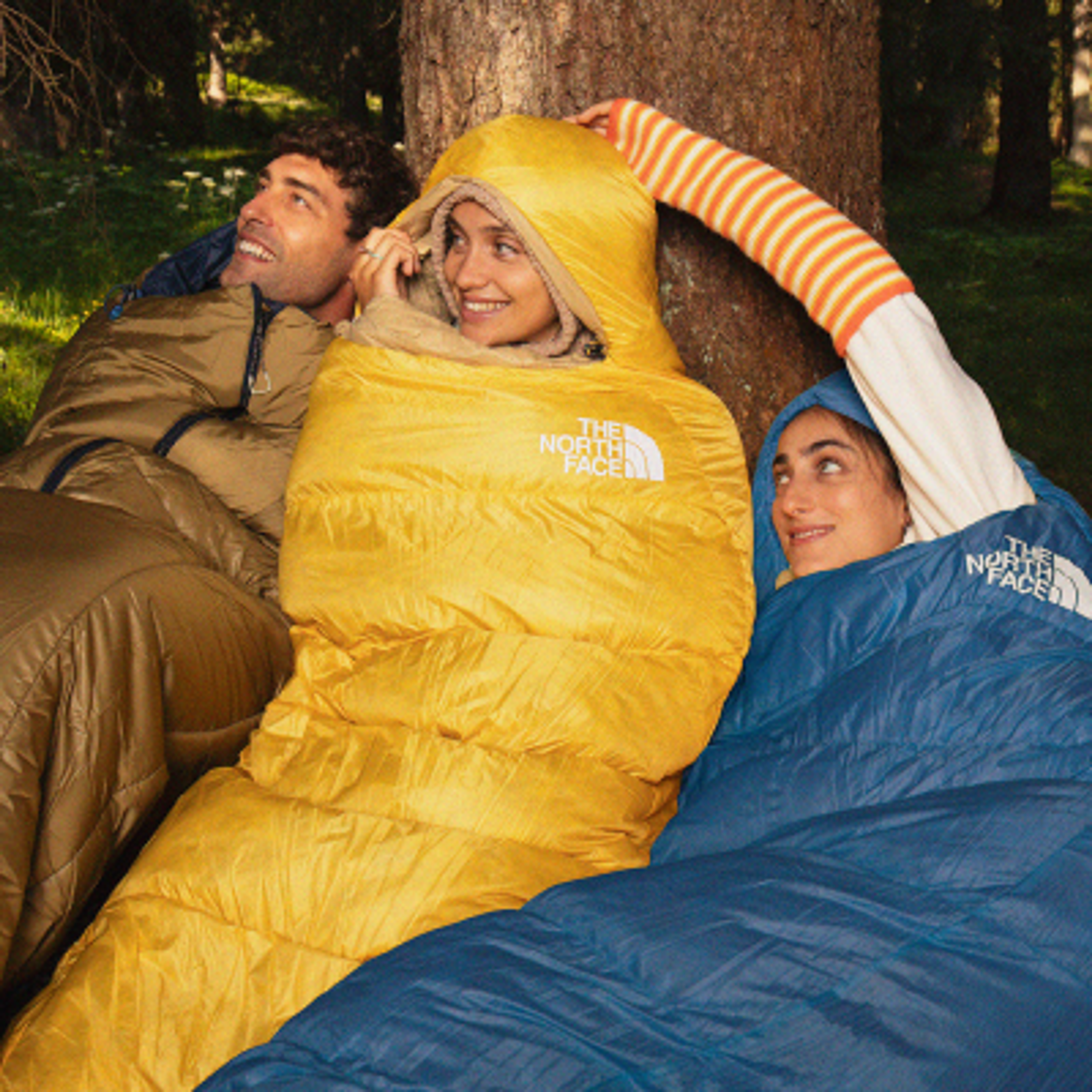 Three people smiling in yellow, blue, and brown The North Face sleeping bags by a tree.