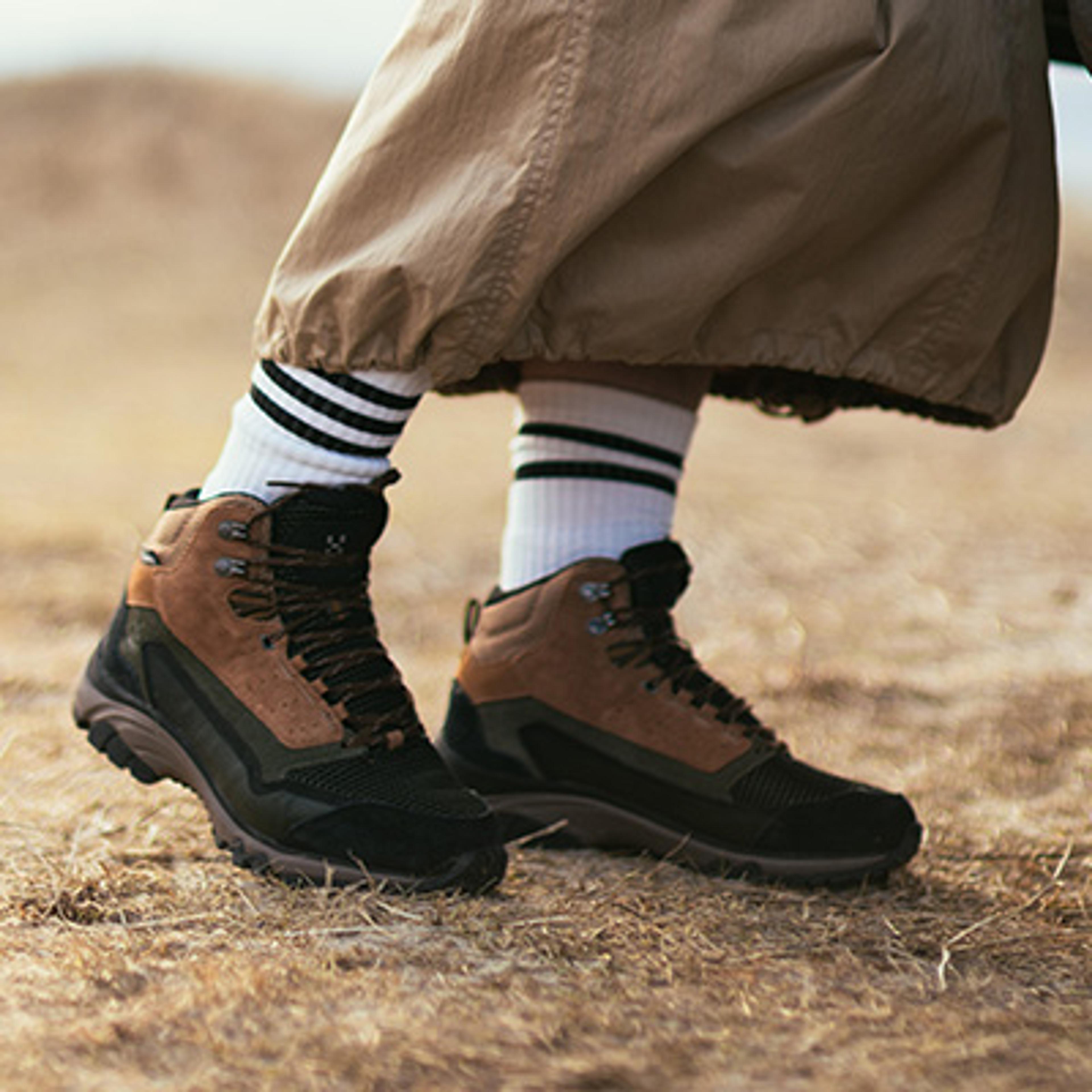 Brown, green, and black hiking boots, white striped socks, and tan pants on dry ground.