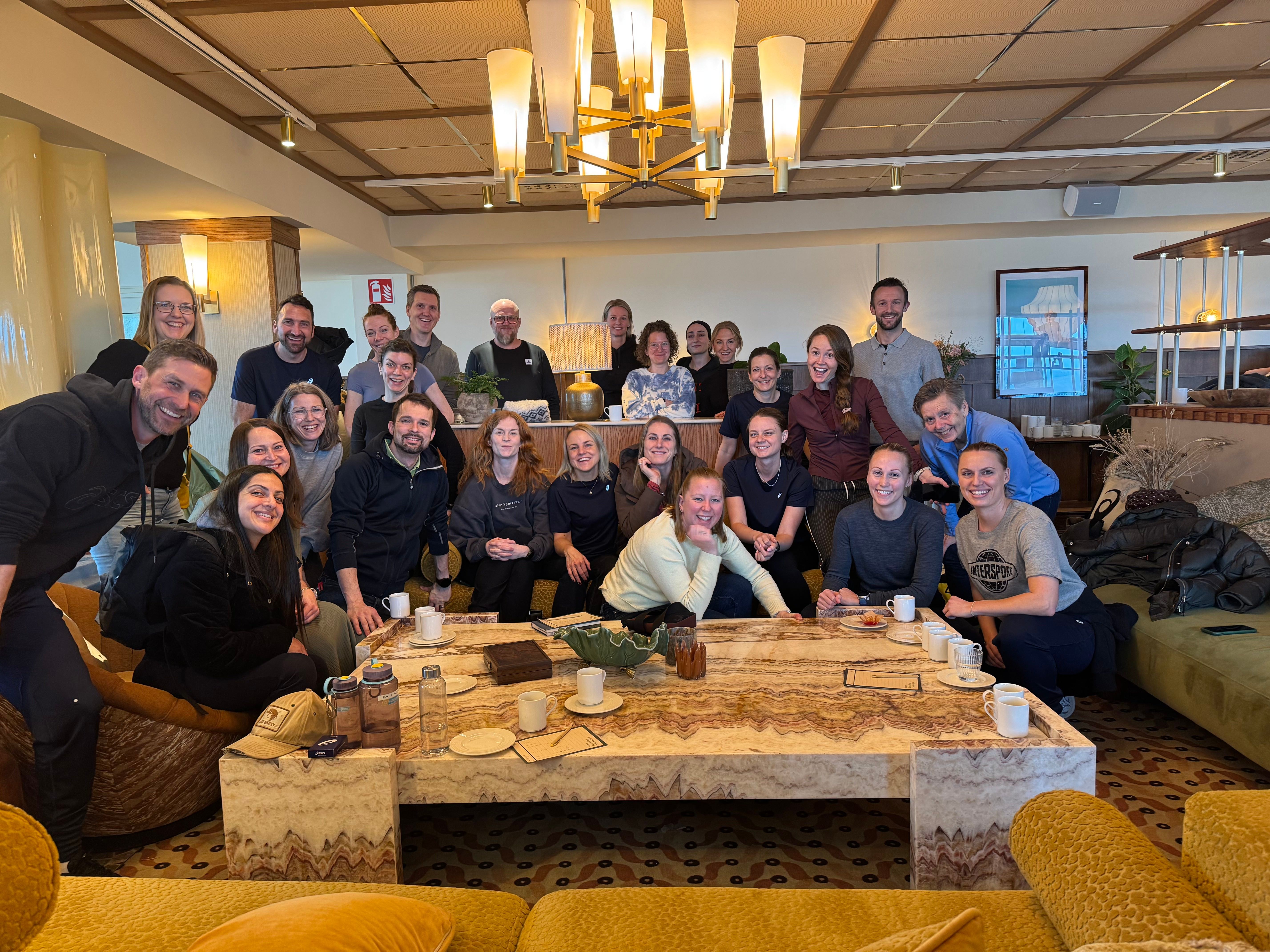 A large group of smiling people gathered in a lounge around a coffee table.