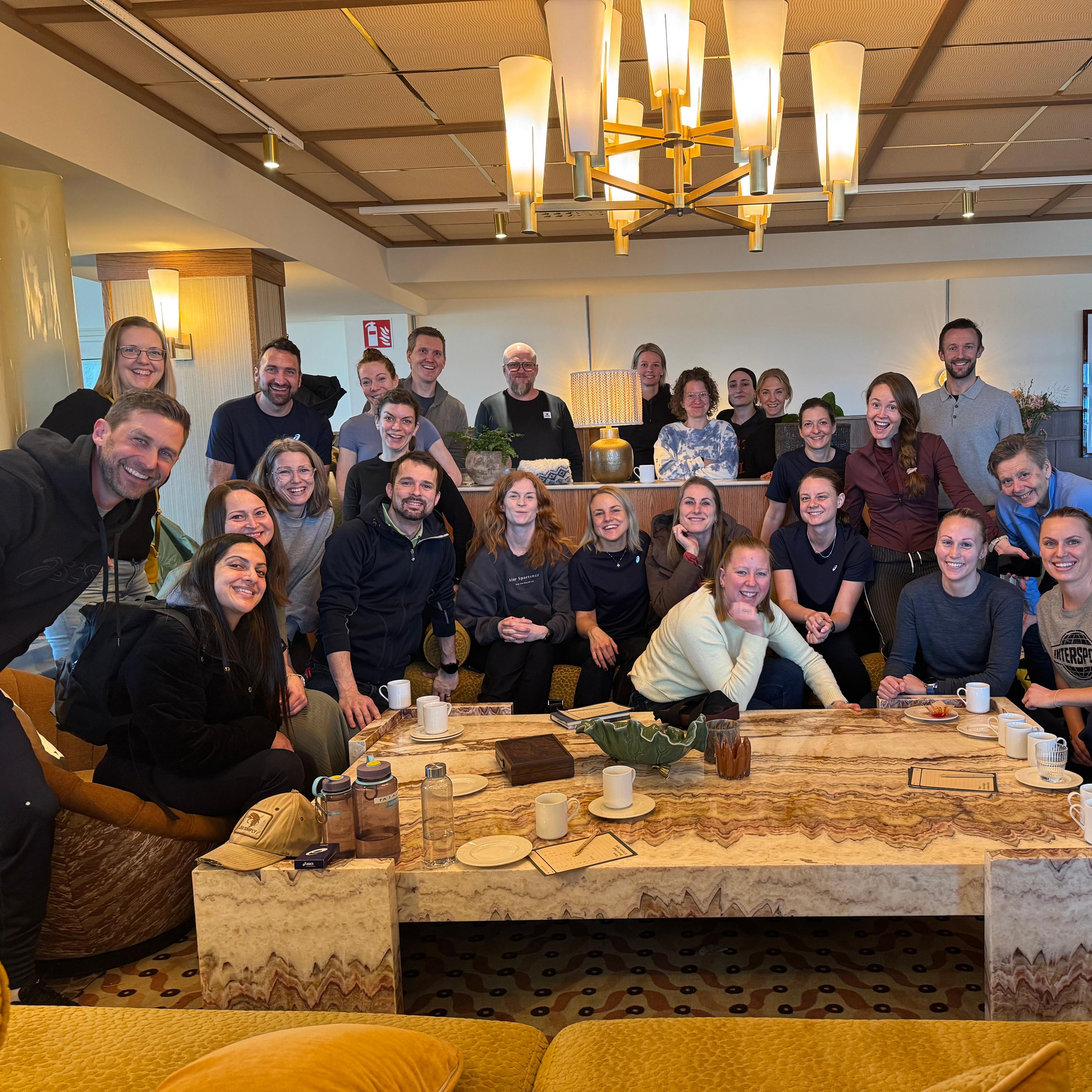 A large group of smiling people gathered in a lounge around a coffee table.
