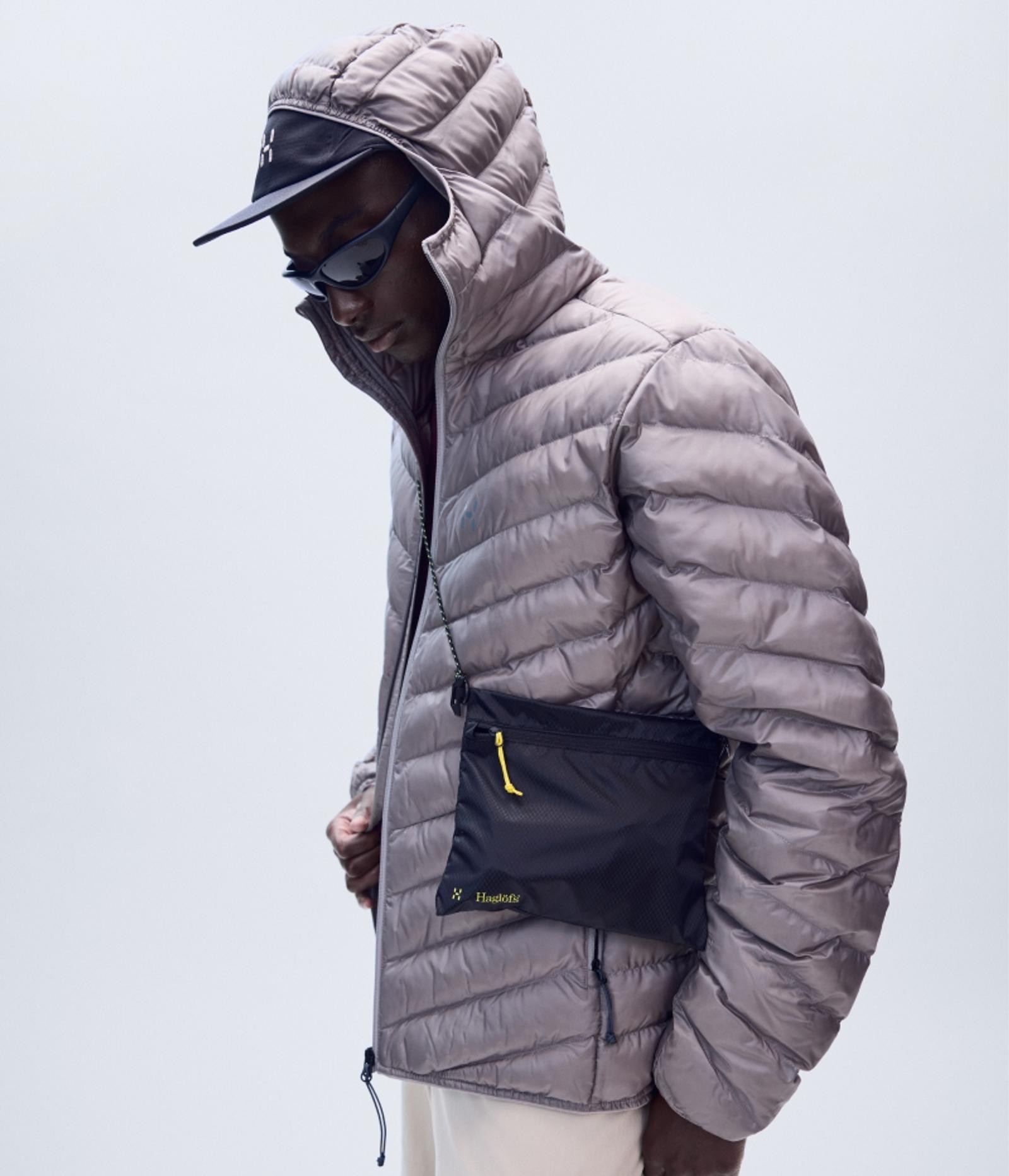 Man in a grey hooded puffer jacket, black cap, sunglasses, and a black crossbody bag.