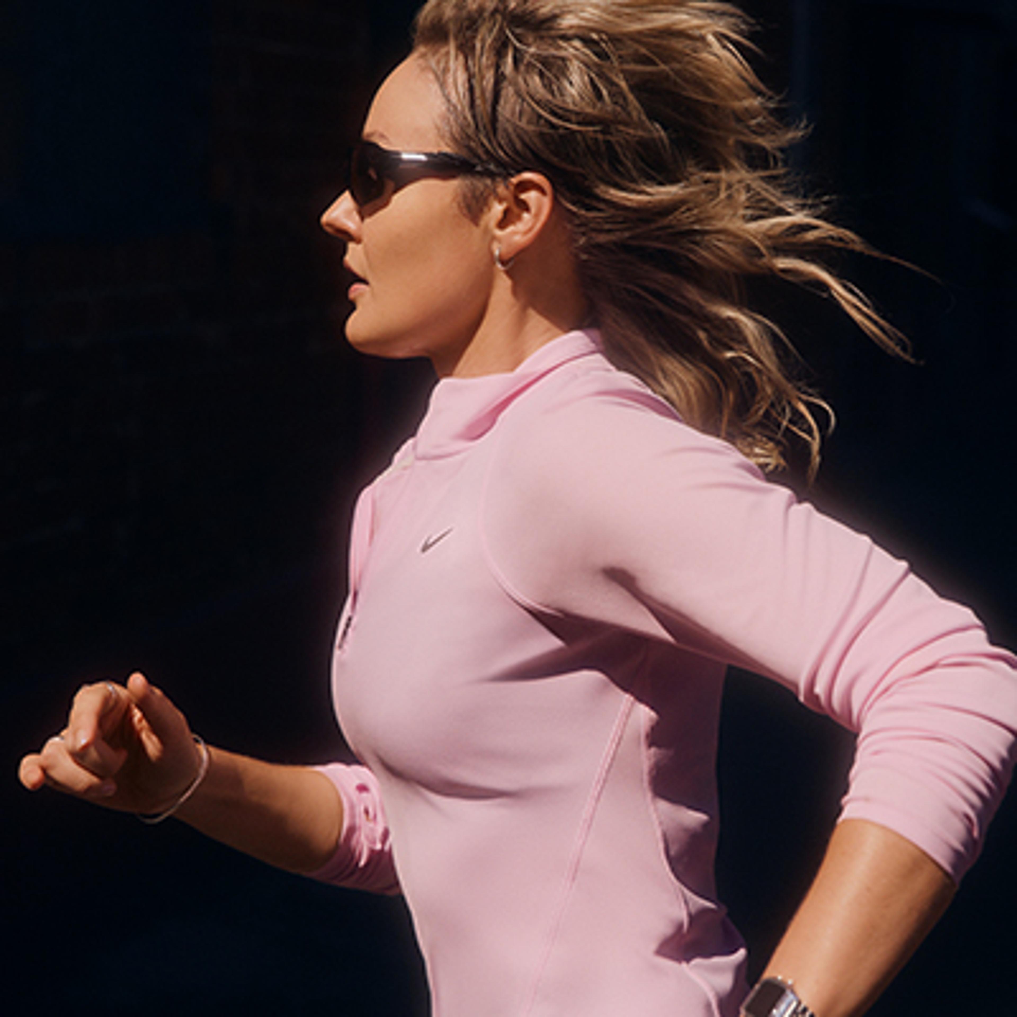 A woman running in profile, wearing sunglasses and a pink Nike athletic top, with her hair blowing back.