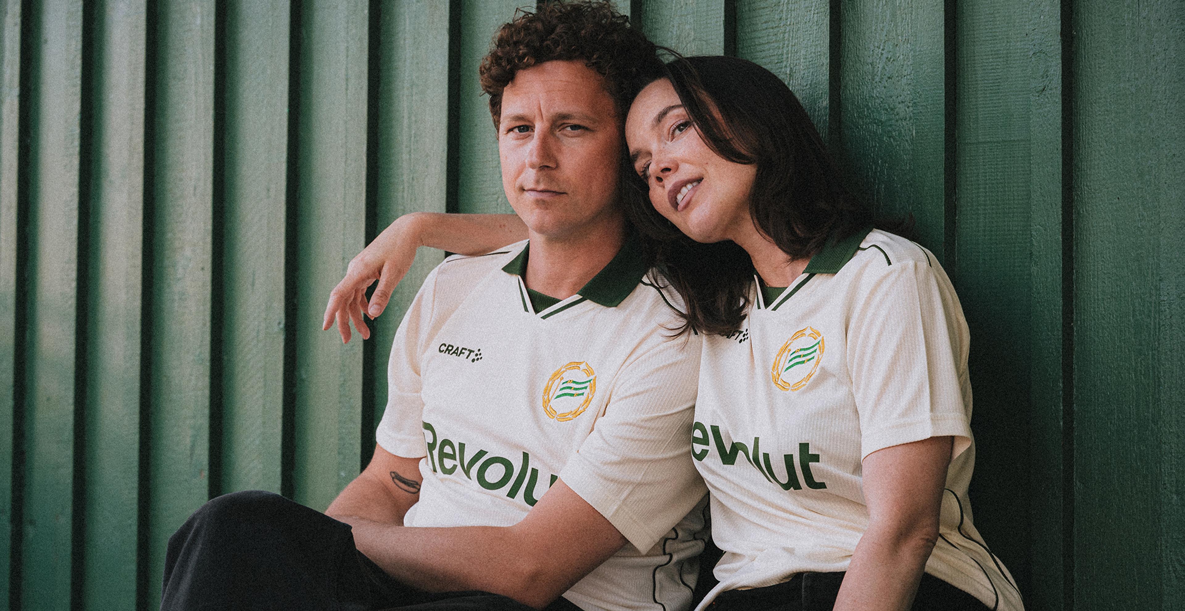 A man and woman in matching cream and green "Revolut" soccer jerseys sit, with the woman's head on the man's shoulder, against a green wooden wall.