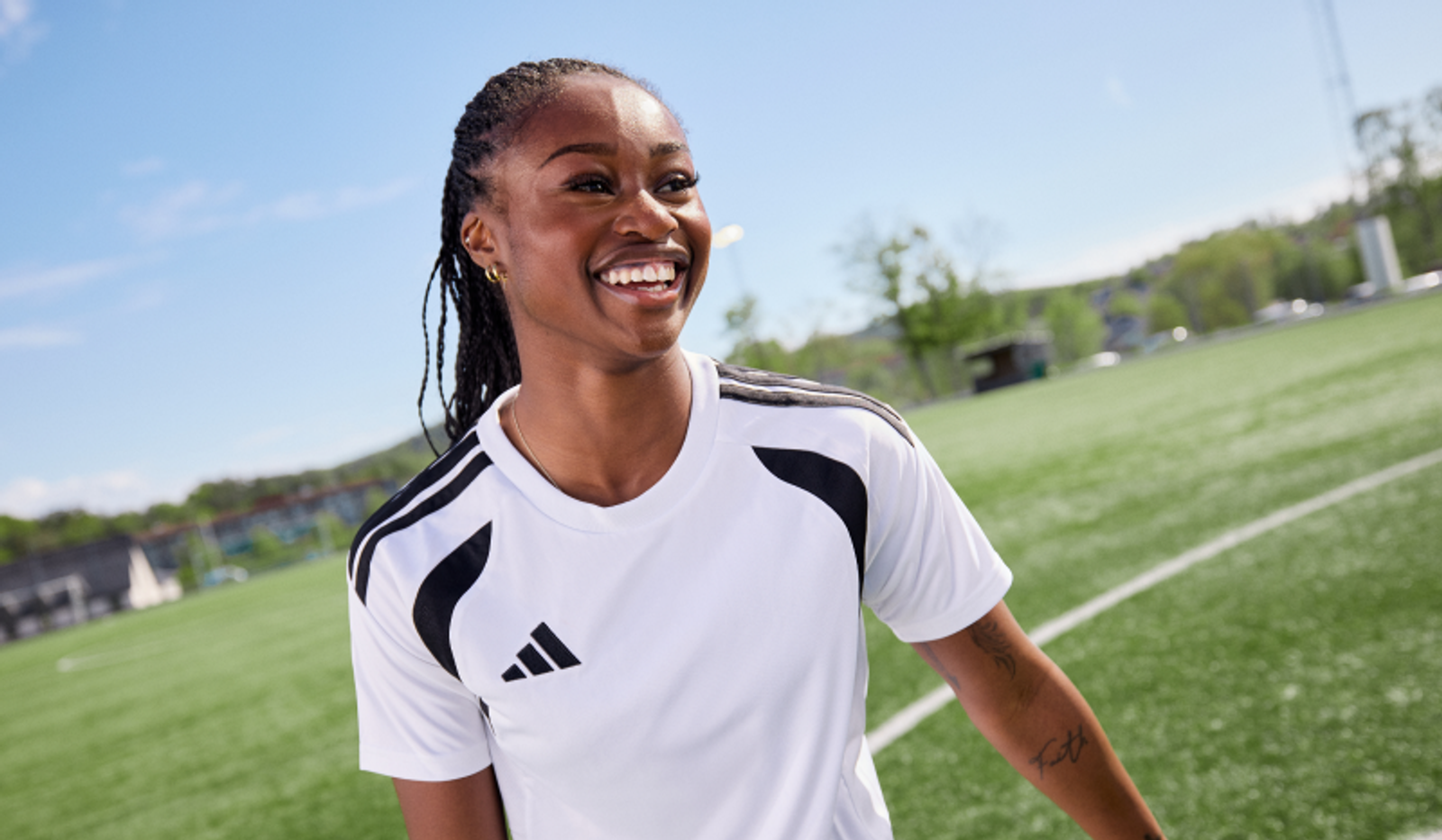 A smiling Black woman in a white Adidas soccer jersey on a green soccer field.
