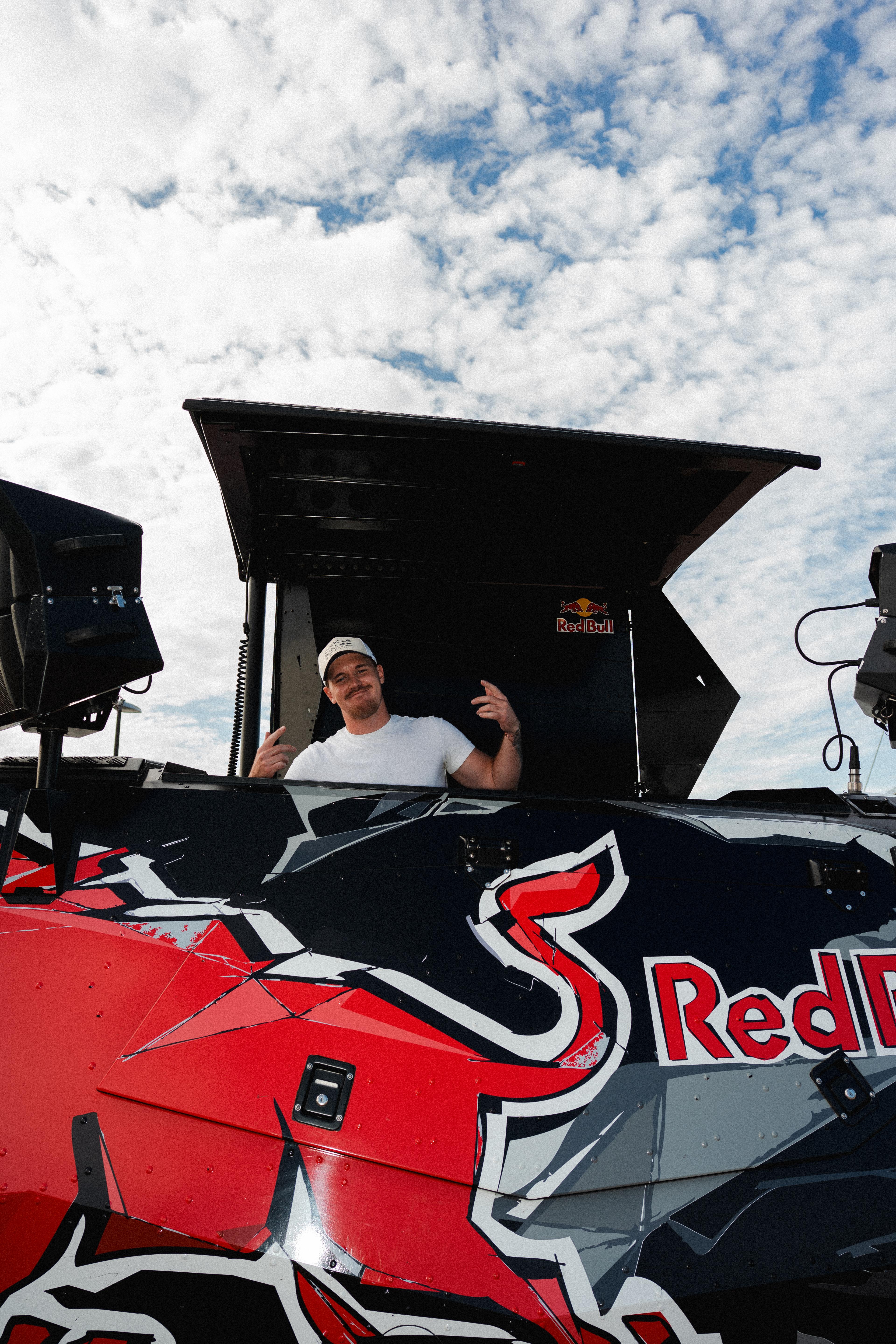 A man in a white cap and t-shirt makes peace signs from a Red Bull branded vehicle against a cloudy sky.