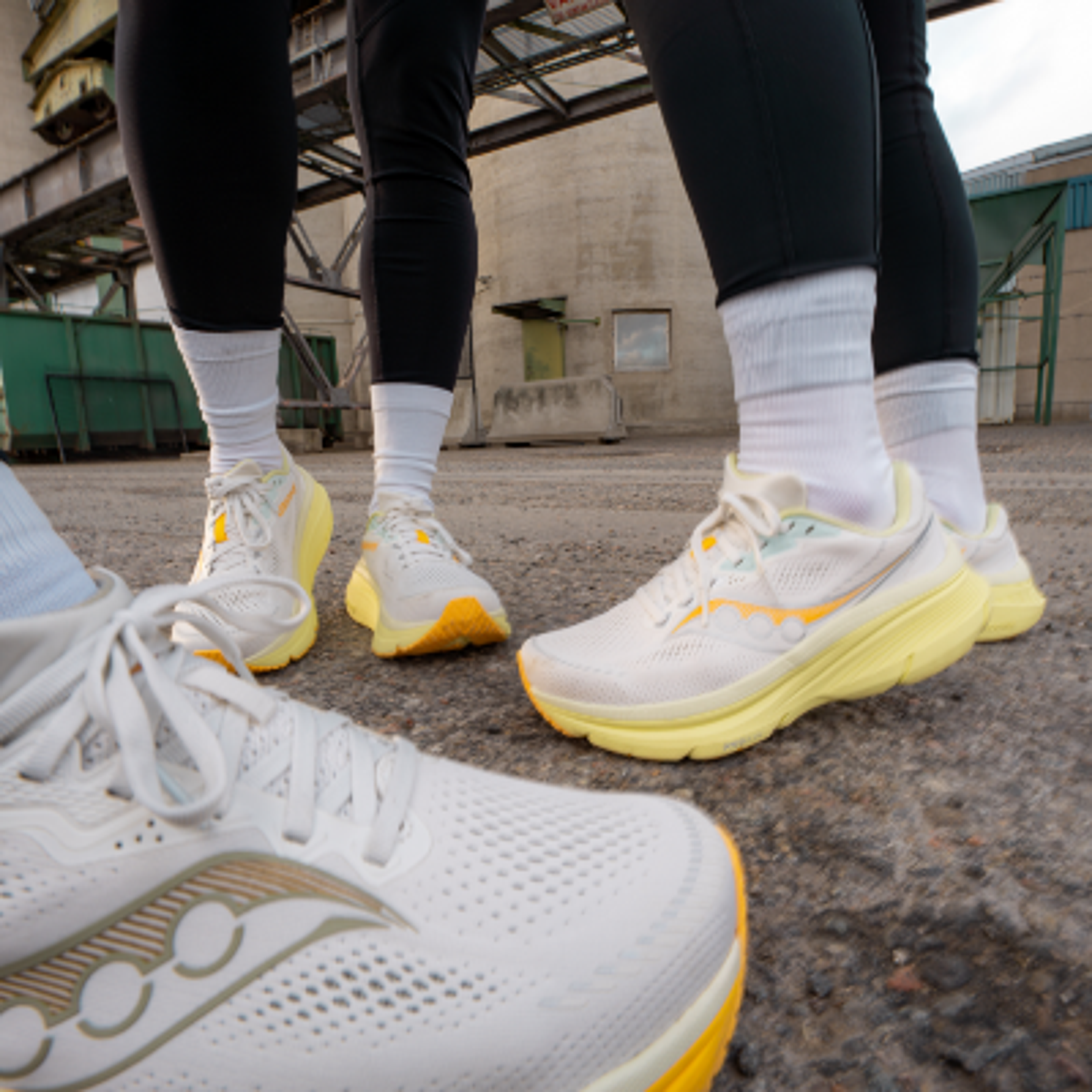 Three pairs of white and yellow athletic shoes on a concrete ground, worn by people in black leggings.