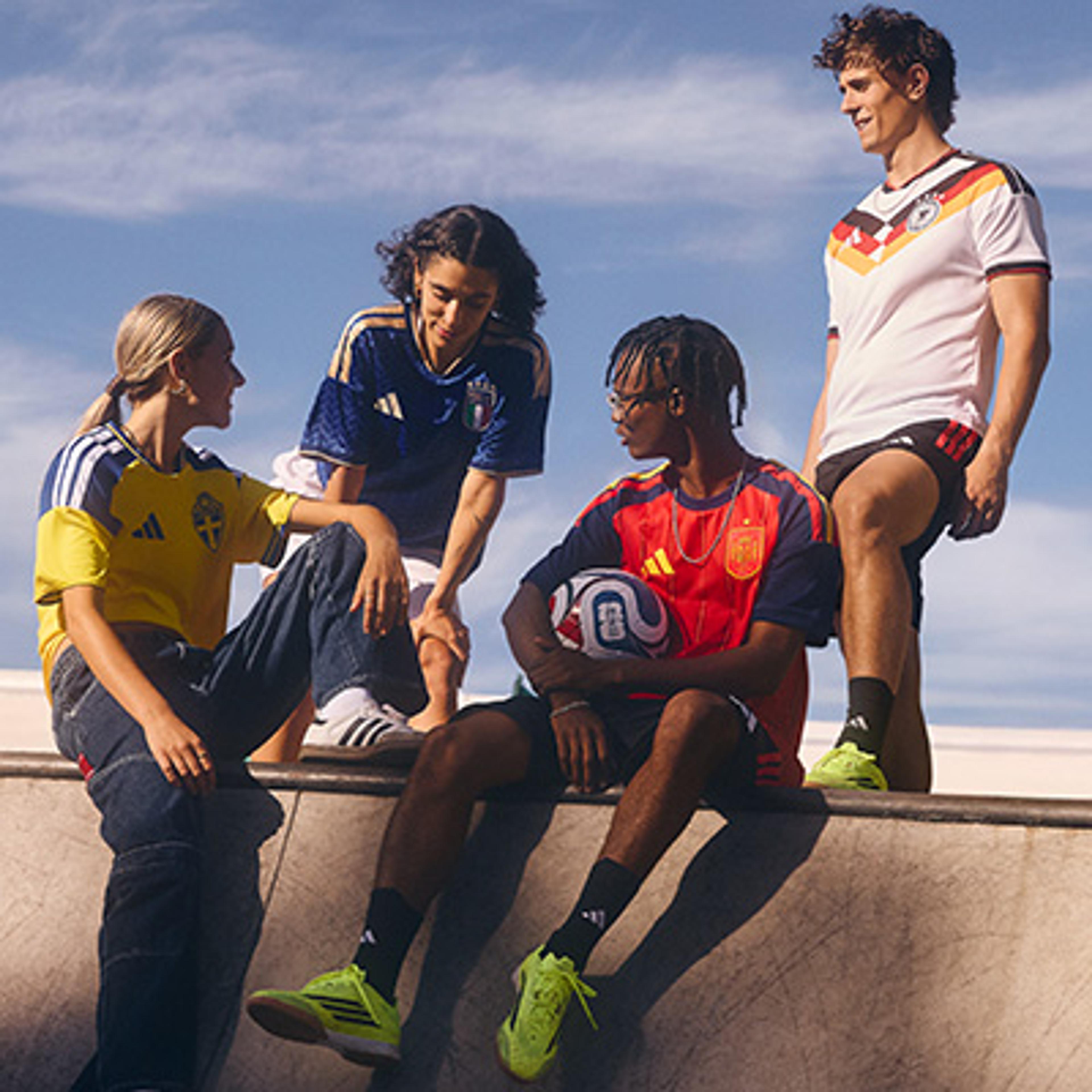 Four young adults wearing Sweden, Italy, Spain, and Germany national soccer jerseys, one holding a ball, in an outdoor urban setting.