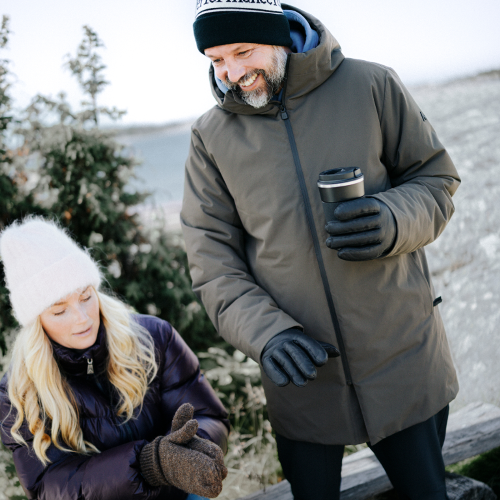 A smiling man in a winter coat and beanie holds a thermos while looking at a woman in a white beanie and purple coat, rubbing her gloved hands together.