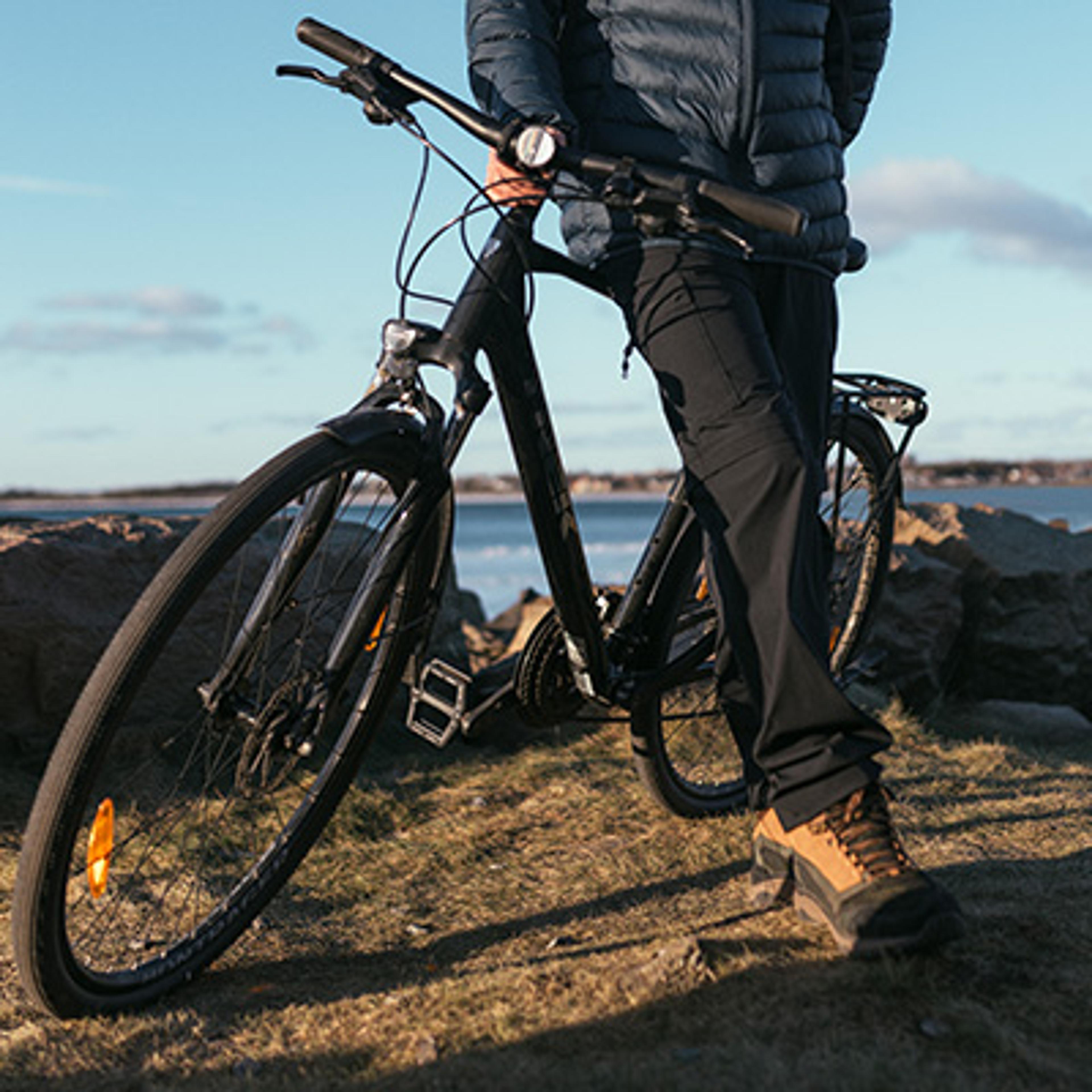 A person stands with a black bicycle on a rocky shore overlooking the sea.