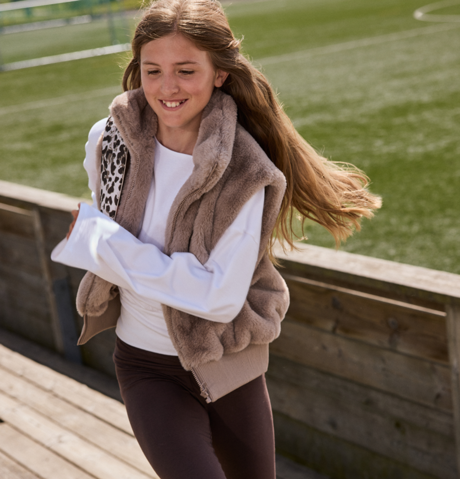 A smiling girl in a brown faux fur vest and white shirt, with her long hair blowing, outdoors.
