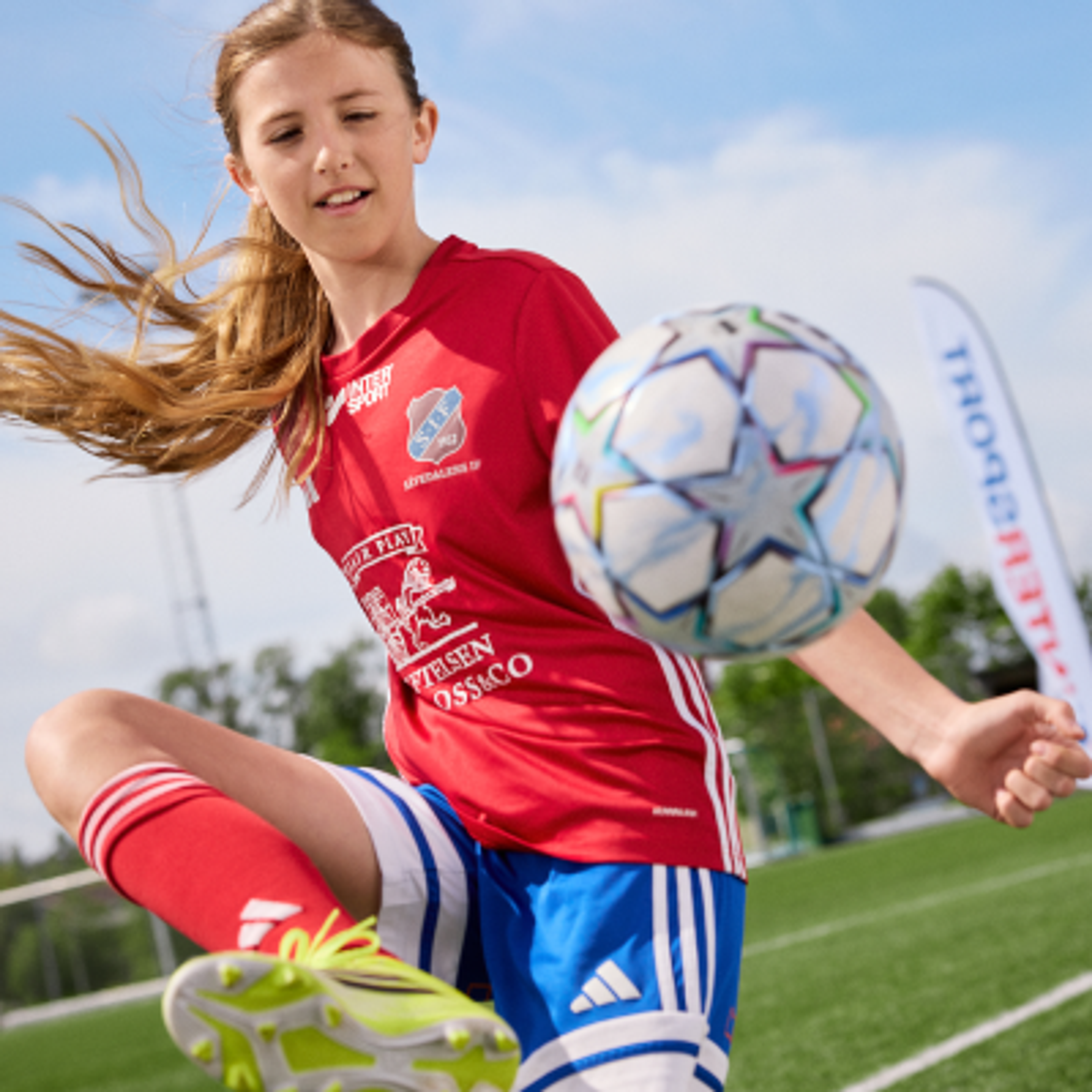 Young girl in a red soccer jersey kicking a ball on a green field.