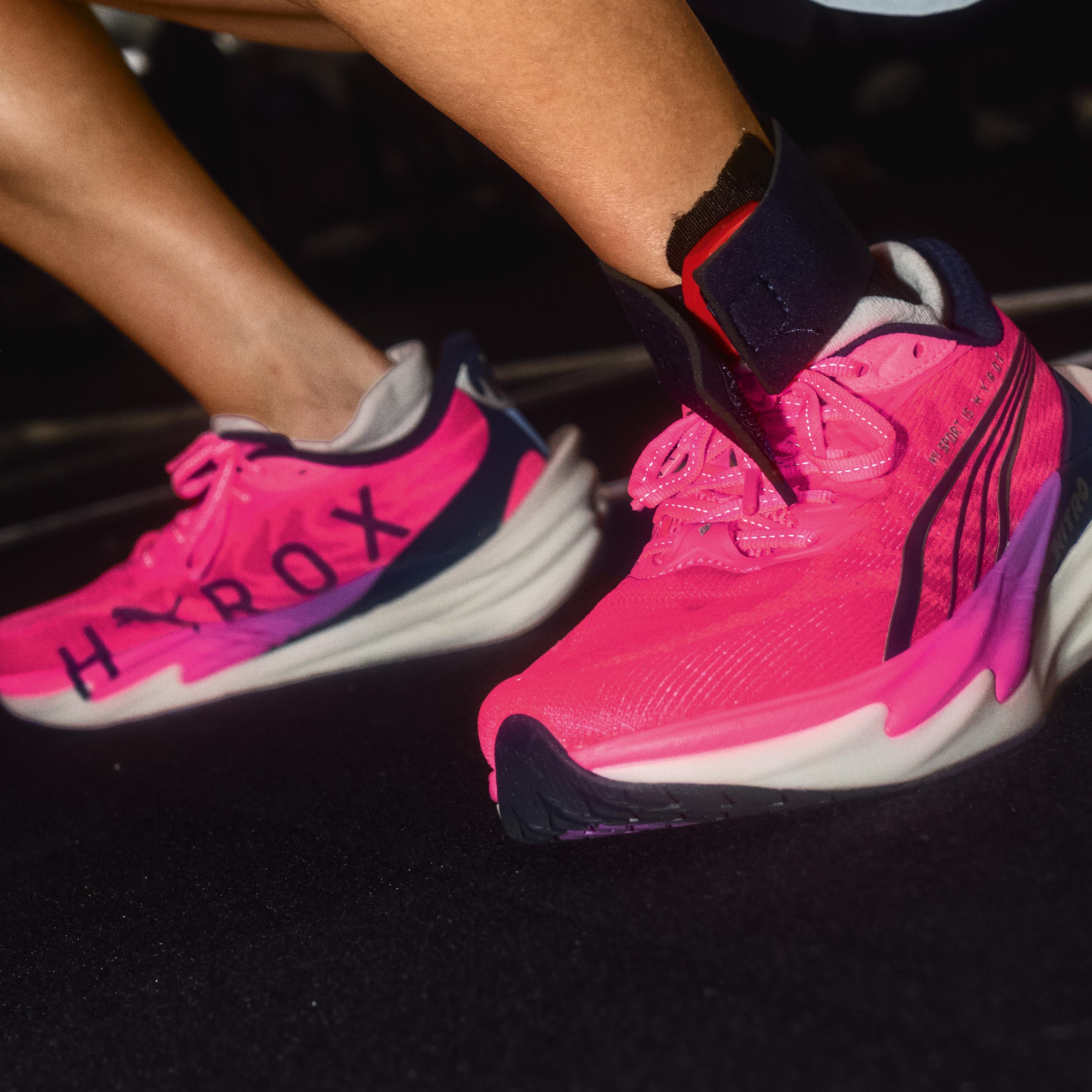 A woman in a light blue top and pink shoes squats, holding a black and grey medicine ball close to her face.