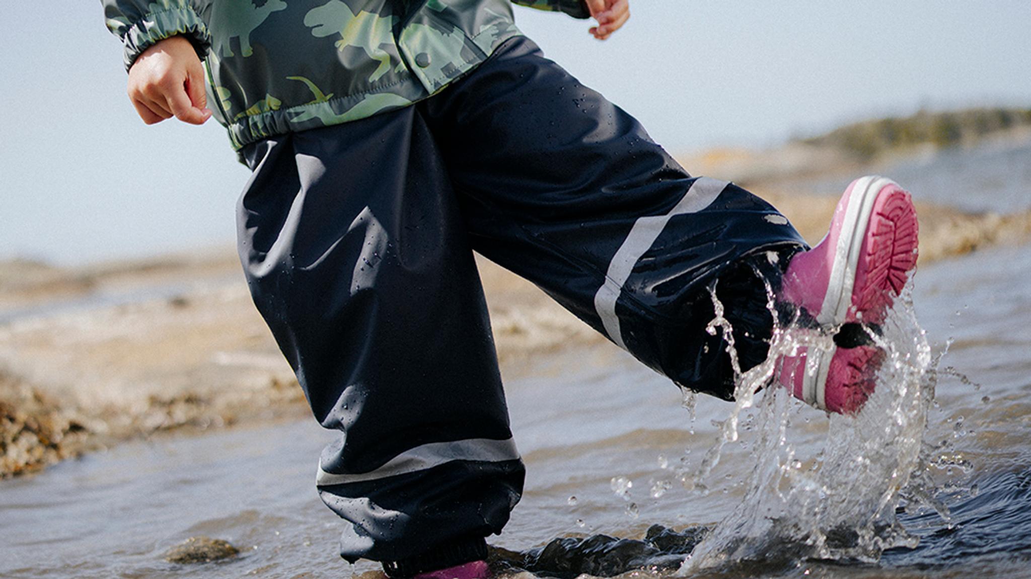 A child in a green rain jacket, dark waterproof pants, and pink boots splashes water.