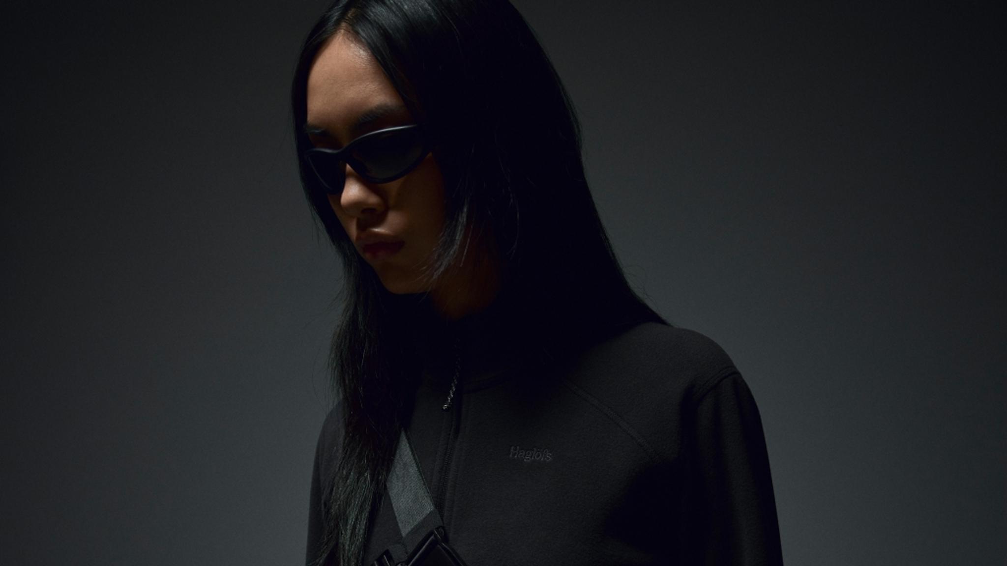 A person with long dark hair, wearing dark sunglasses, a black top, and a cross-body bag.
