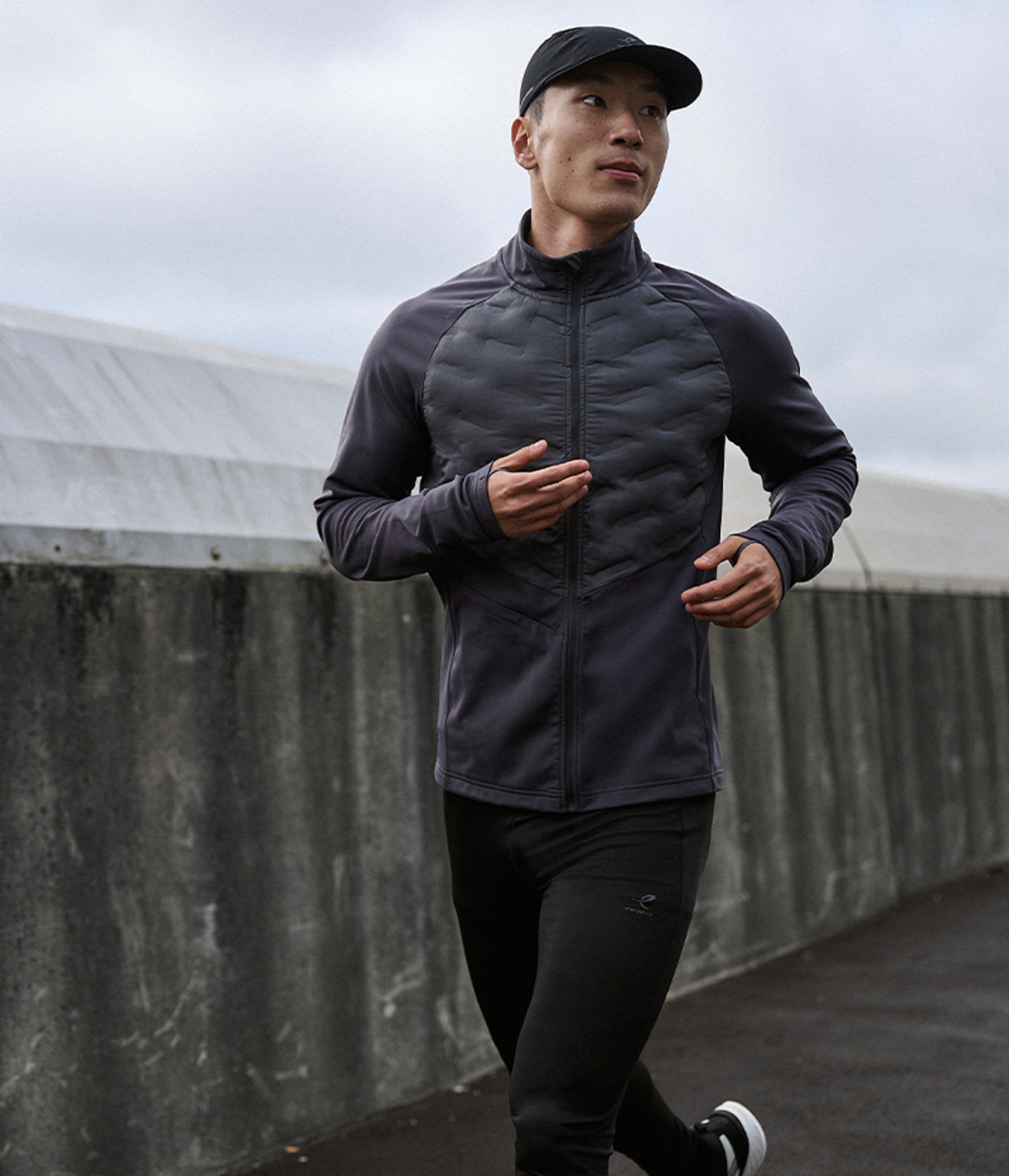 A man in a black cap and dark athletic jacket with a quilted front jogs outdoors.