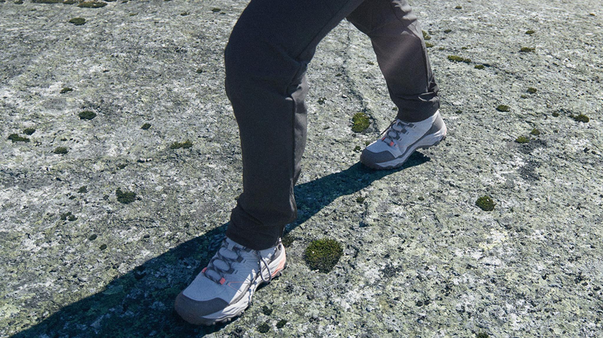 A person's feet in grey hiking shoes walking on lichen-covered rock with green moss.