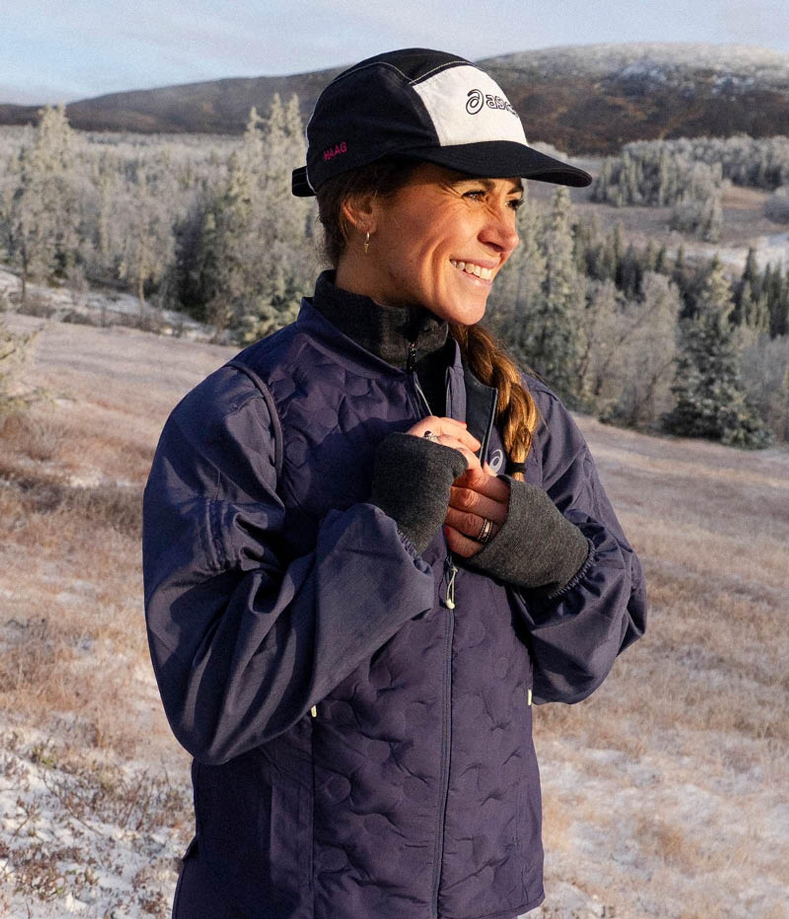 A smiling woman in a navy quilted jacket and cap stands in a snowy, forested landscape.
