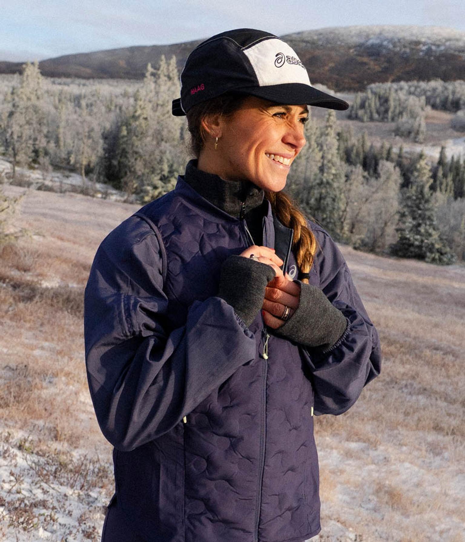 A smiling woman in a navy quilted jacket and cap stands in a snowy, forested landscape.
