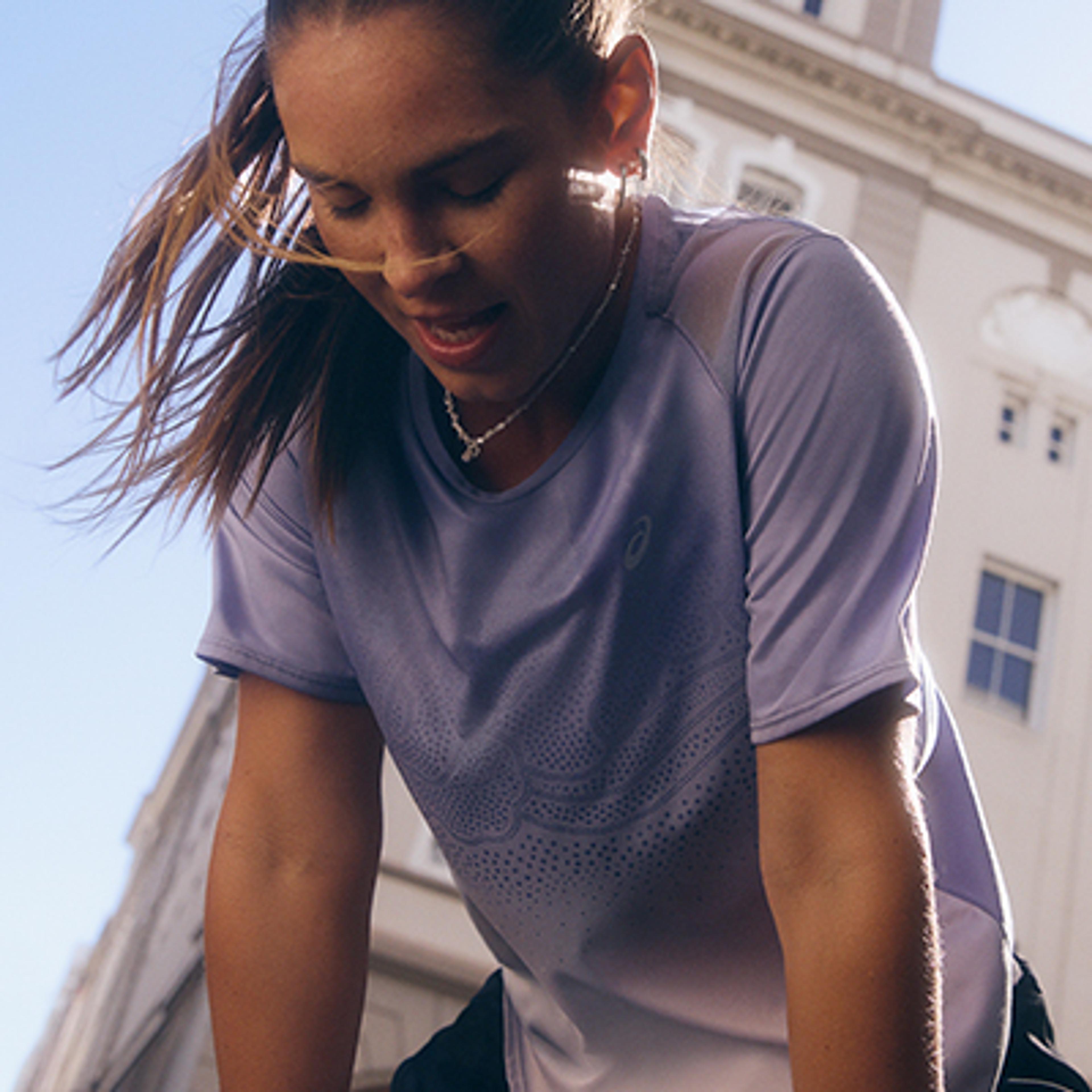 A female runner in a light purple ASICS shirt, sweating and looking down to catch her breath outdoors.