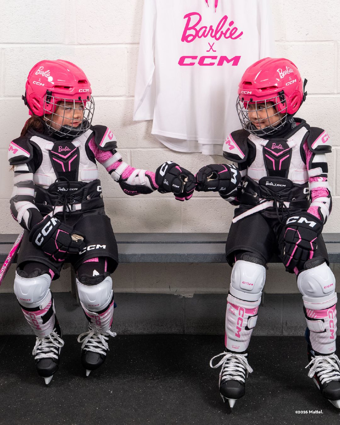 Two girls in pink and black Barbie x CCM hockey gear sit on a bench and fist-bump, with a matching jersey hanging behind them.