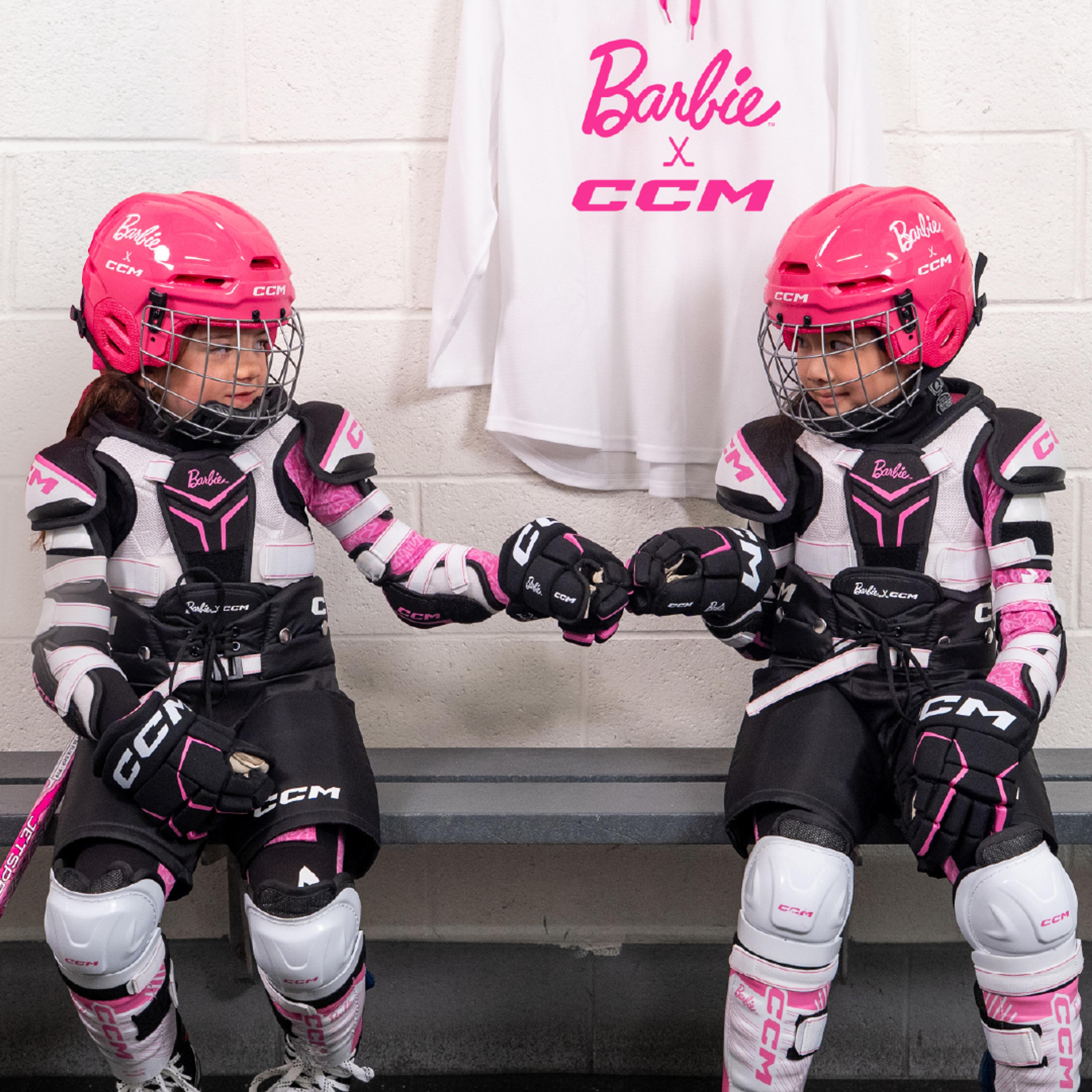 Two girls in pink and black Barbie x CCM hockey gear sit on a bench and fist-bump, with a matching jersey hanging behind them.
