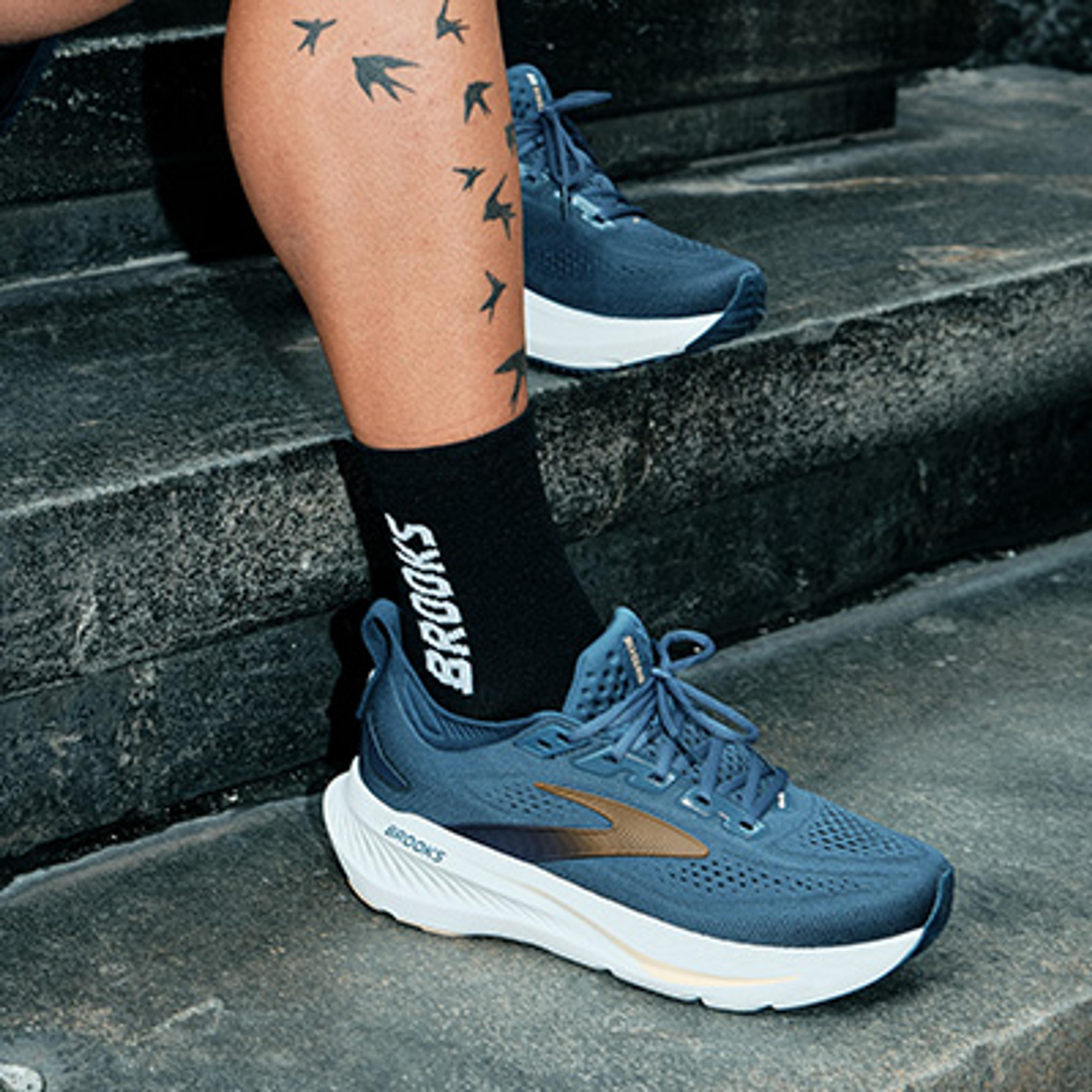 A person's leg with bird tattoos, wearing black Brooks socks and blue Brooks running shoes, stands on stone stairs.