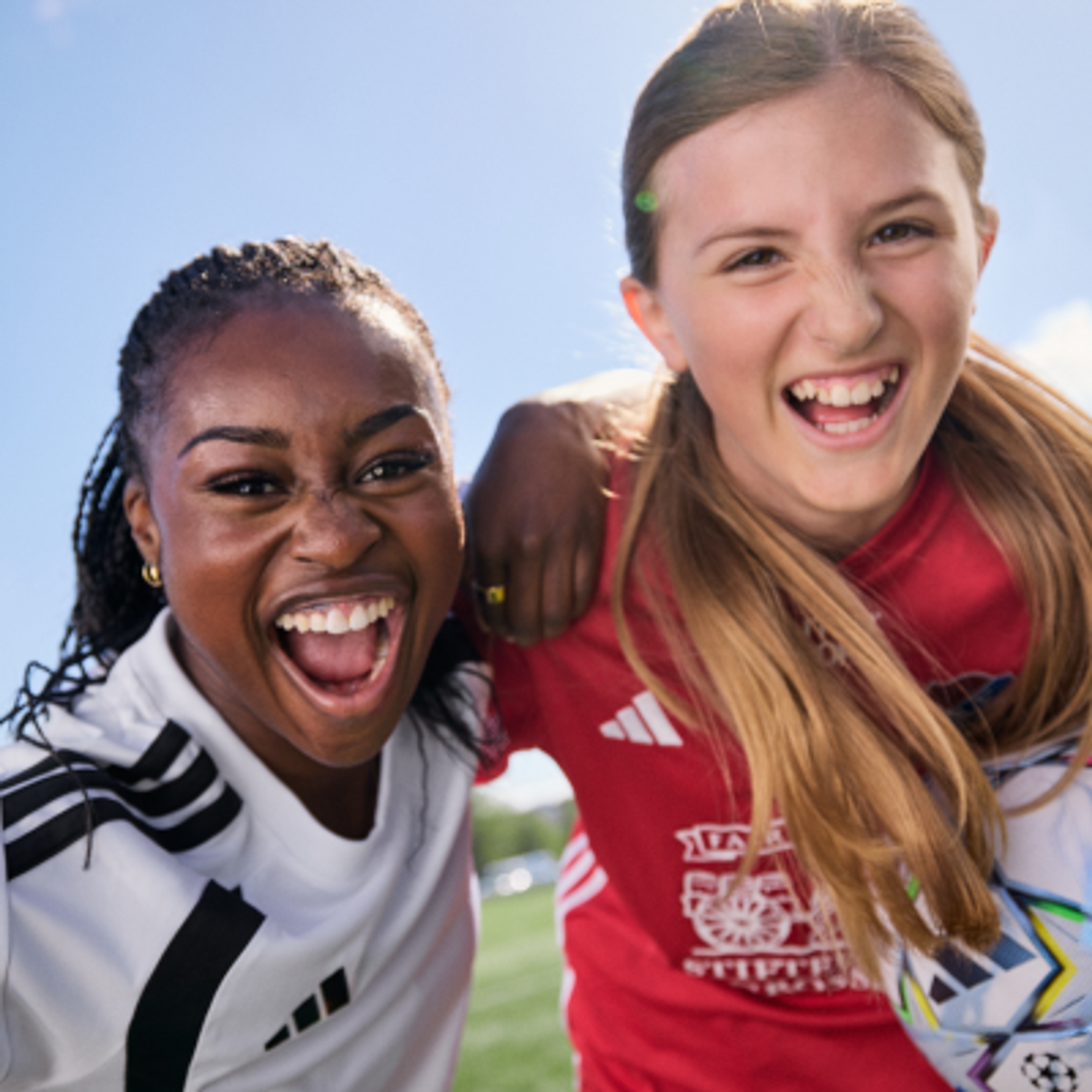 Two smiling young female soccer players, one Black and one White, embrace on a sunny field.