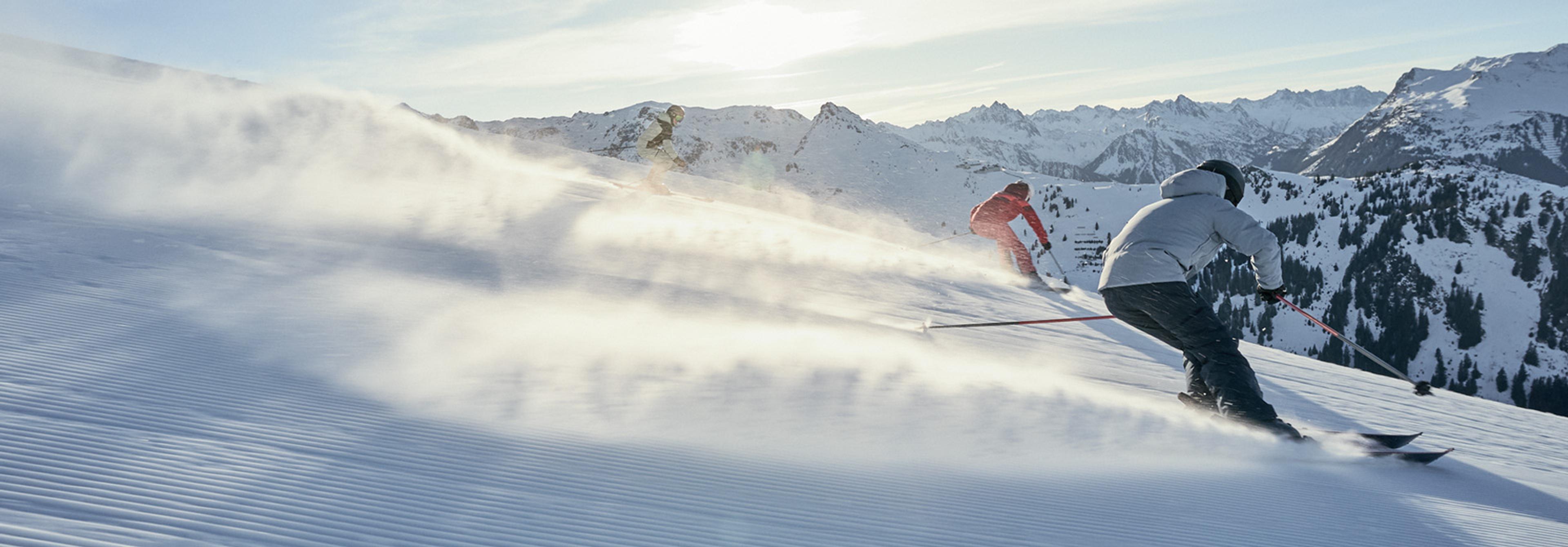 Three skiers descend a sunny mountain slope, kicking up snow.