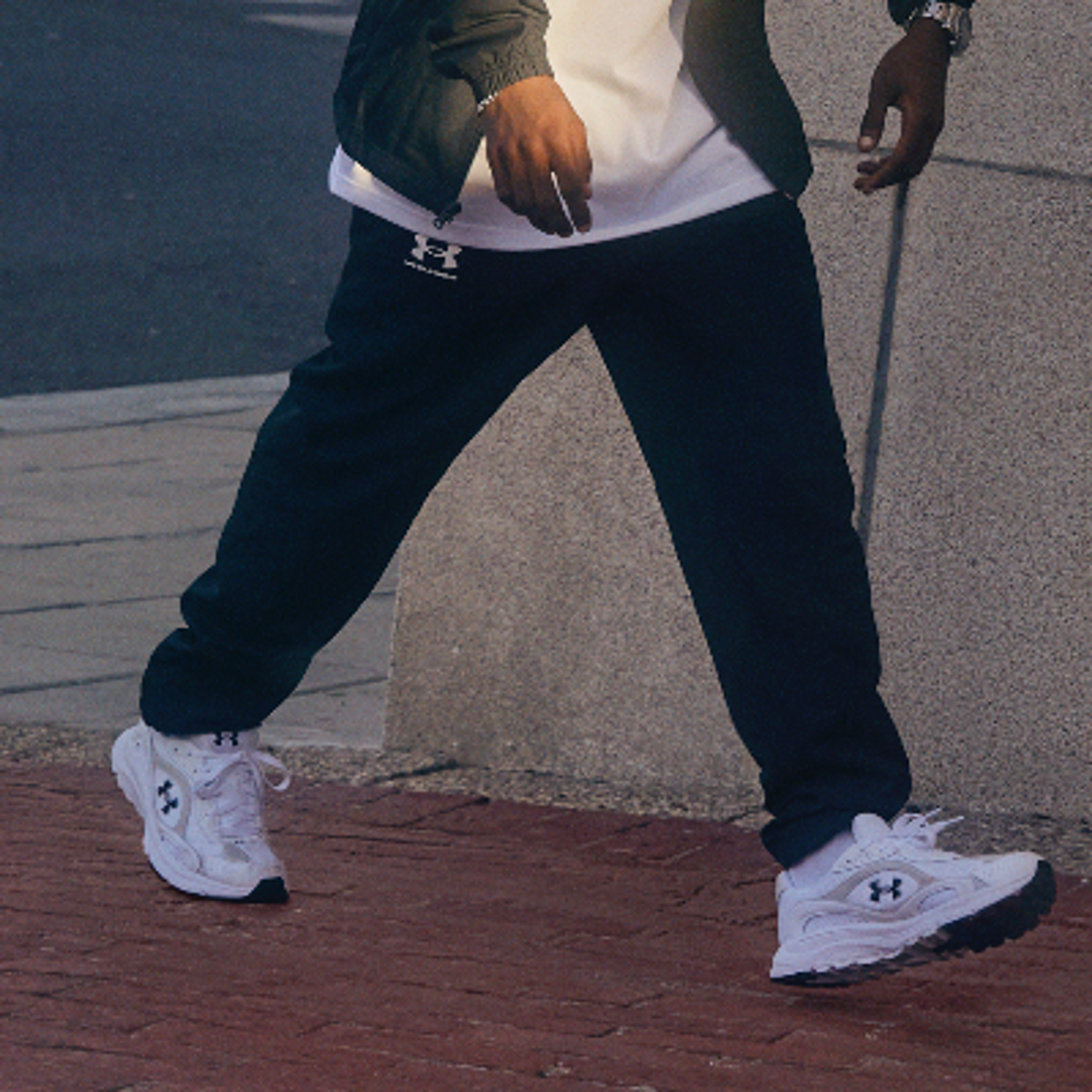 A person wearing dark Under Armour sweatpants and white Under Armour sneakers walks on a brick sidewalk.