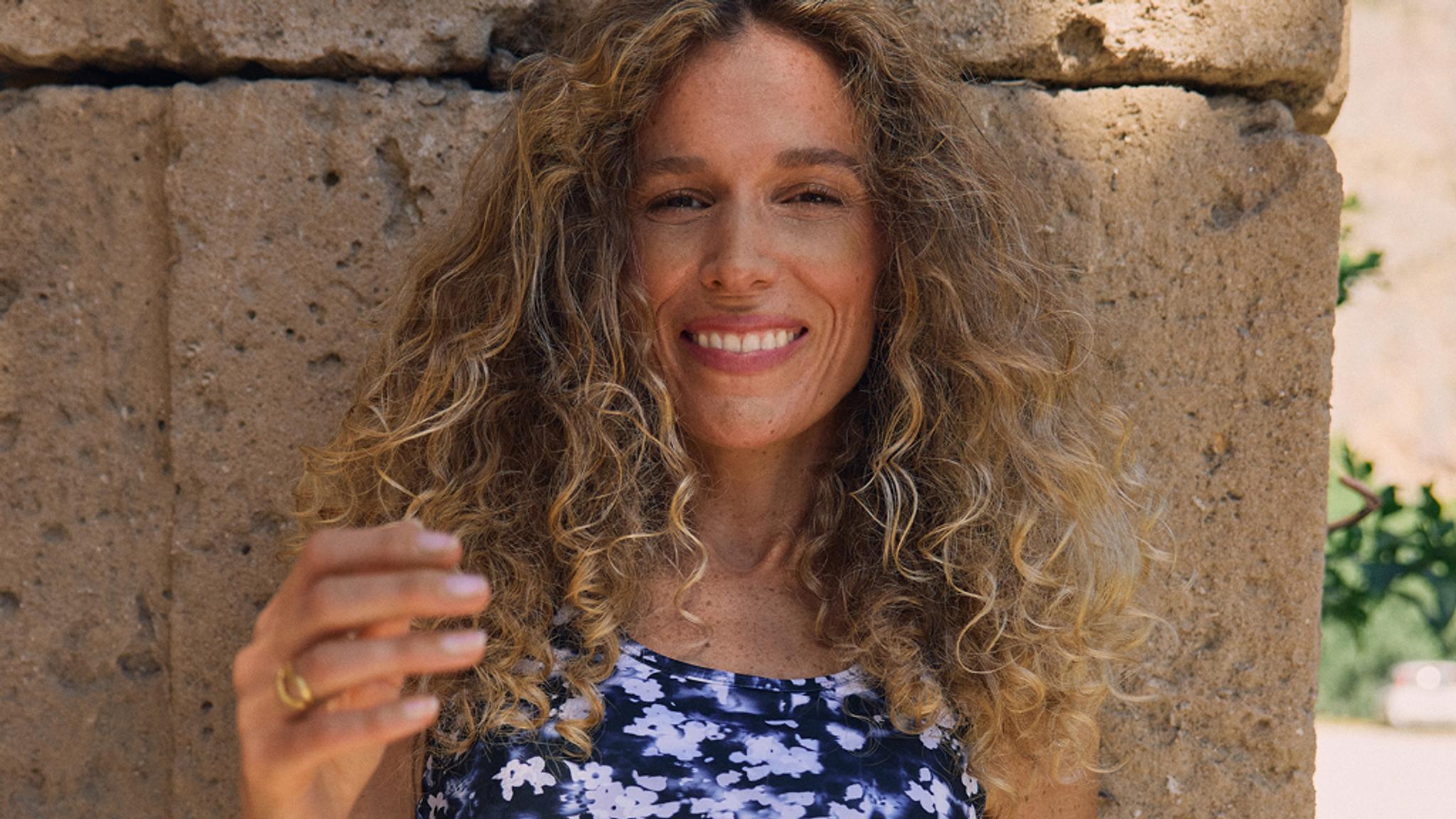 A smiling woman with long curly blonde hair against a stone wall.