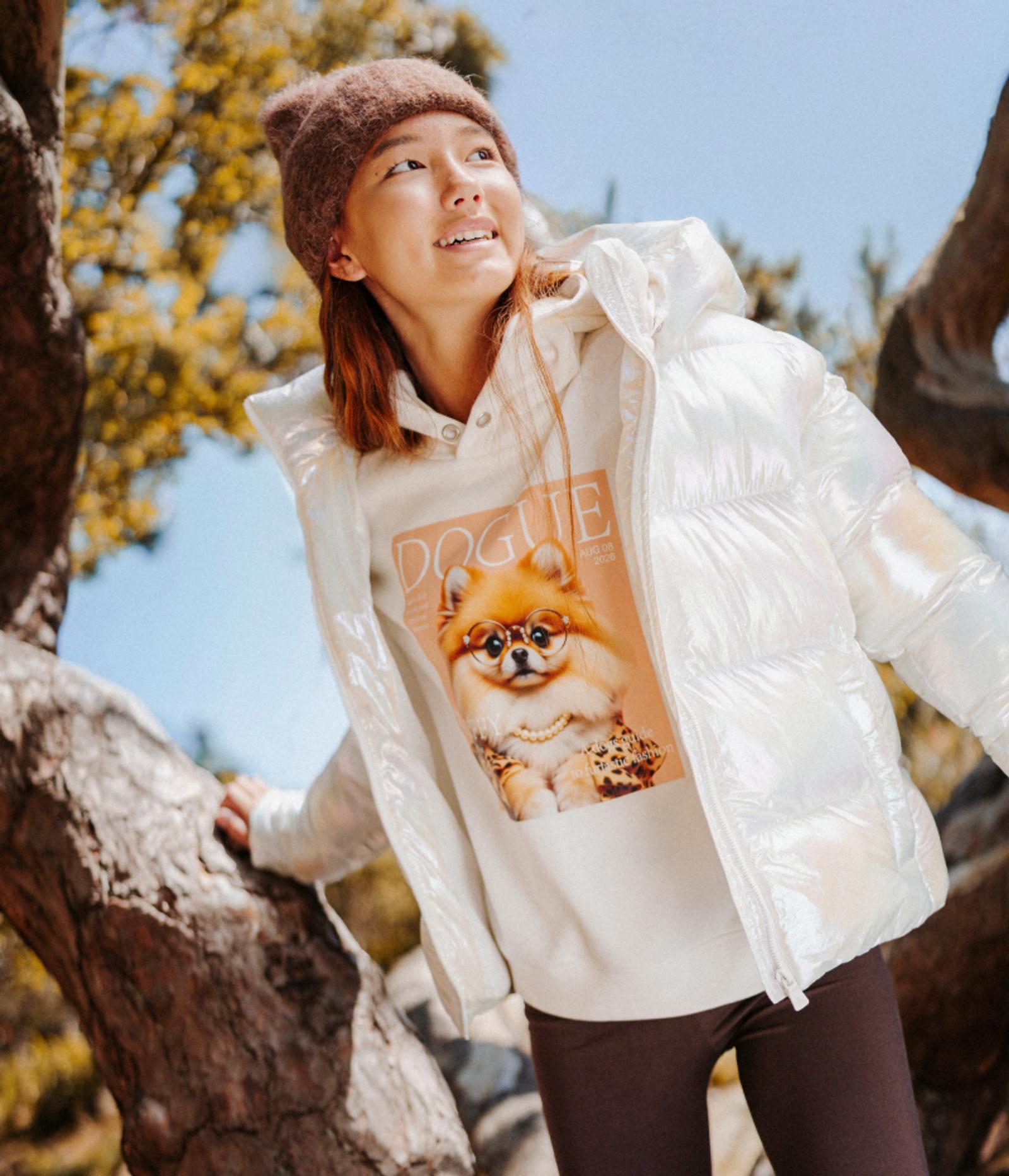 A young person in a brown beanie and iridescent white puffer jacket, wearing a hoodie with a Pomeranian on a "Dogue" magazine cover, smiling upwards outdoors.