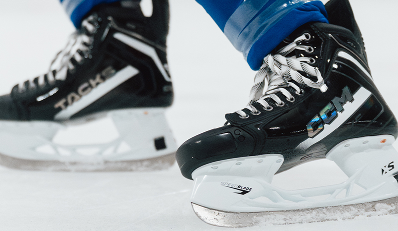 Close-up of two black hockey skates on an ice rink, one branded CCM and the other Tacks.