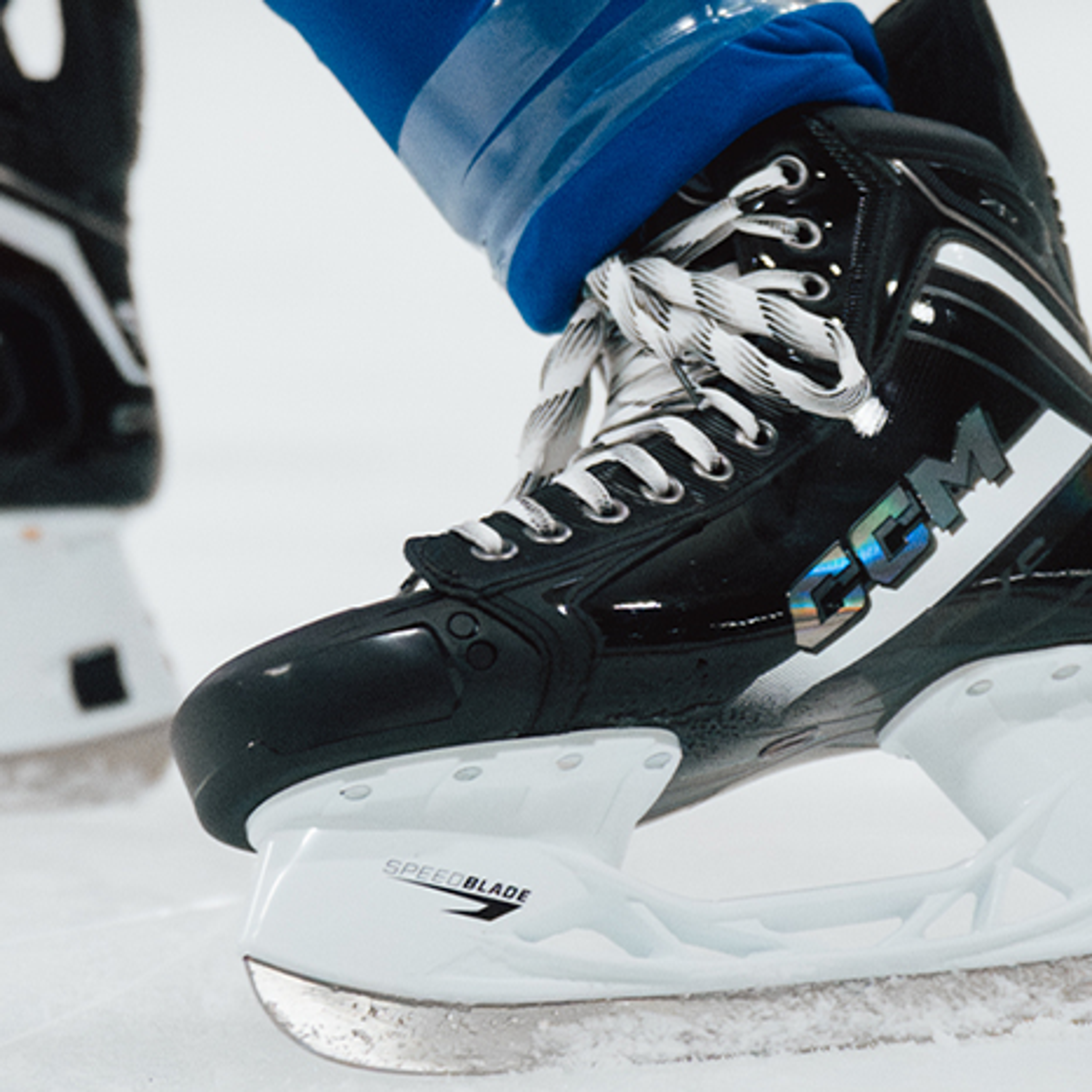 Close-up of two black hockey skates on an ice rink, one branded CCM and the other Tacks.