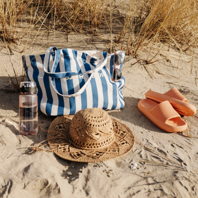 A blue and white striped beach bag, sunglasses, a pink water bottle, a straw hat, and orange slides on sand.