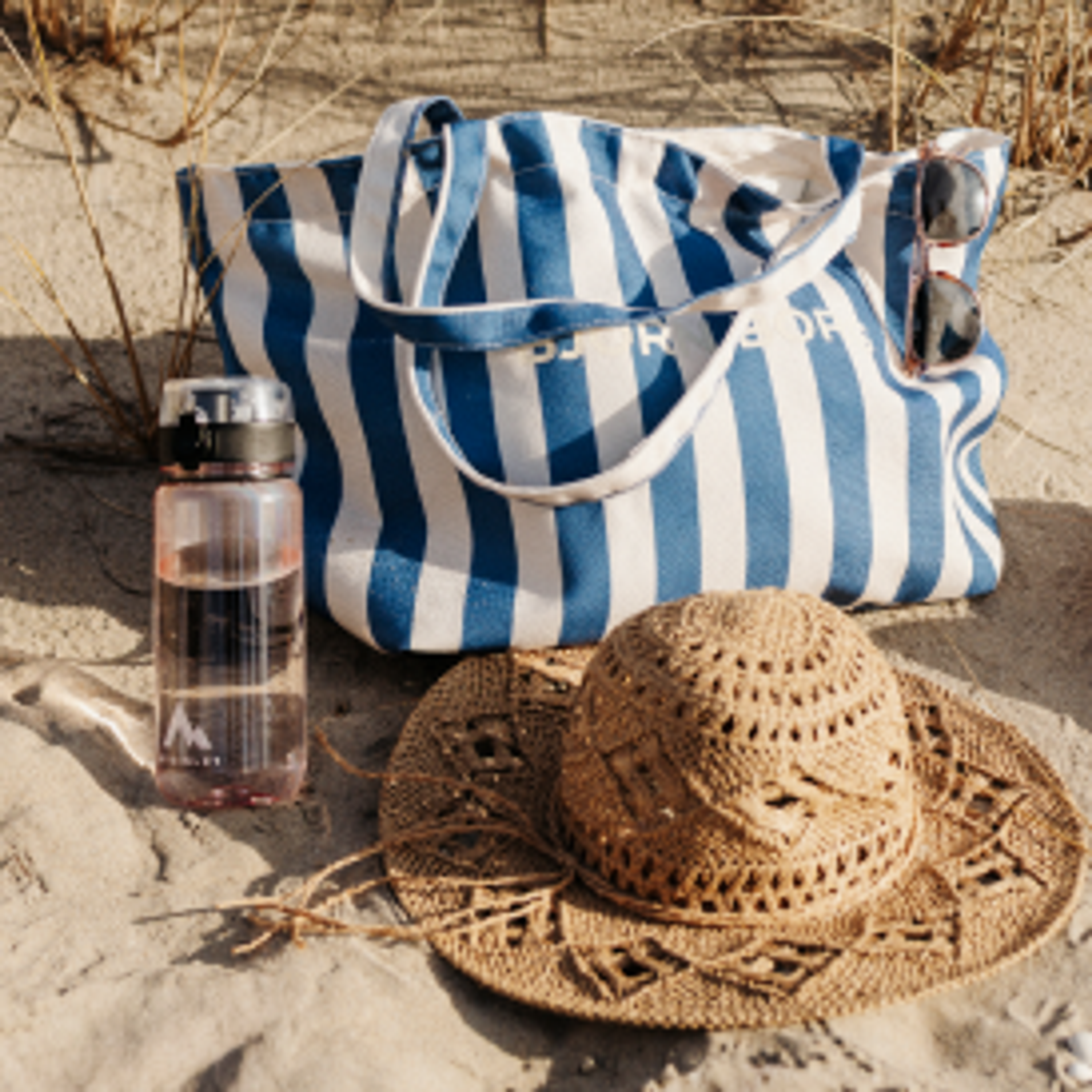 A blue and white striped beach bag, sunglasses, a pink water bottle, a straw hat, and orange slides on sand.