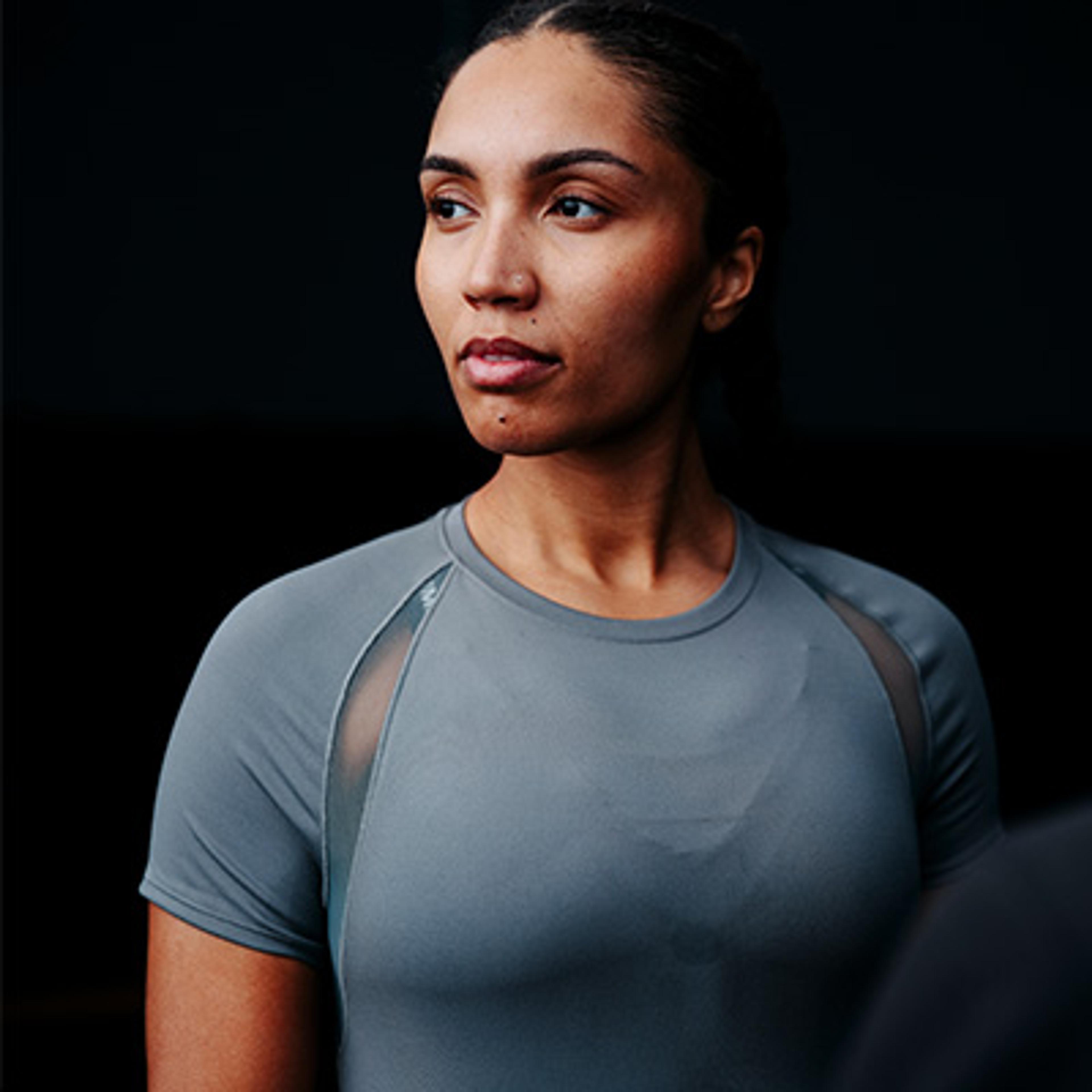 A dark-skinned woman in a grey athletic shirt with mesh panels looks to the side.