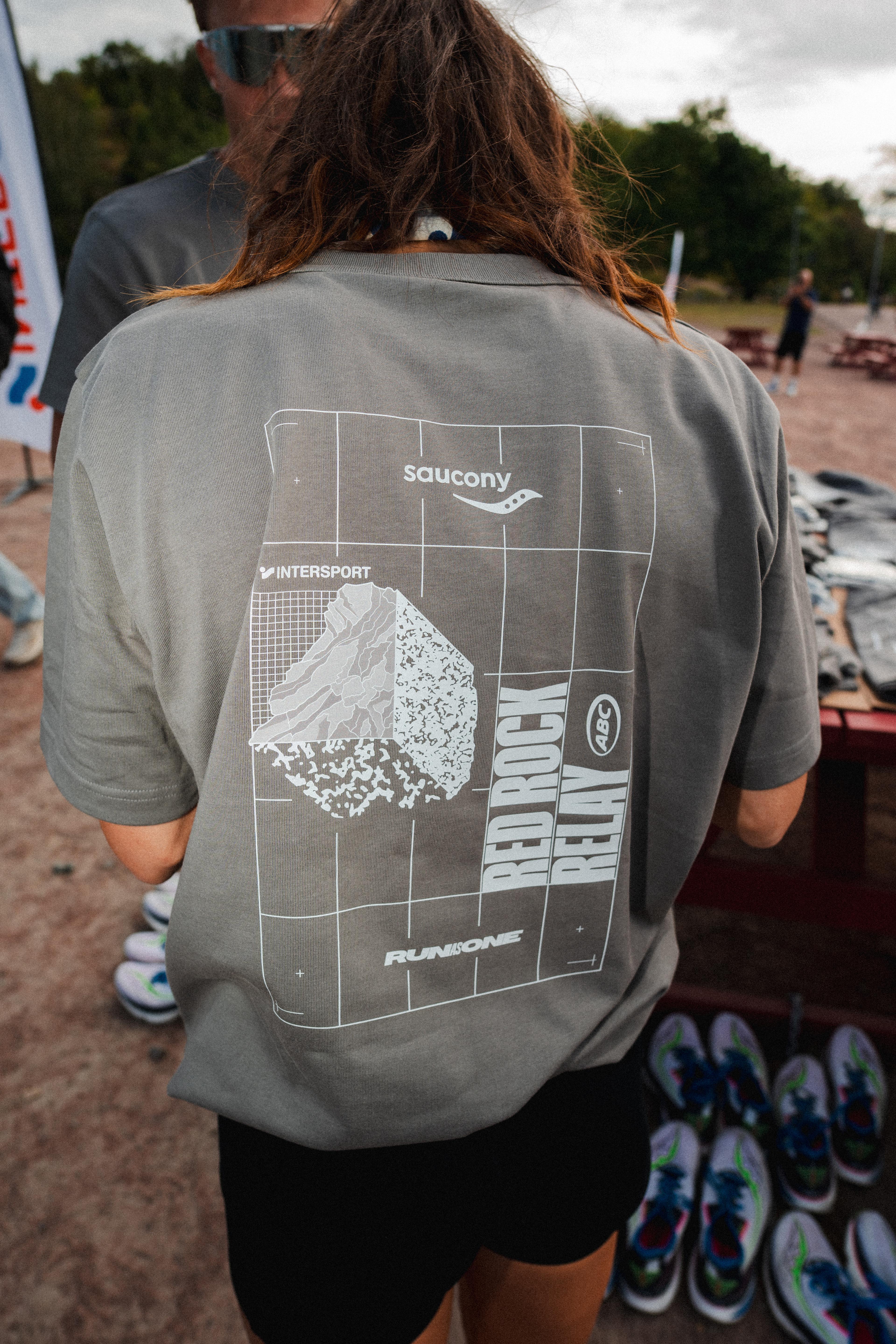A person from behind wears a gray t-shirt featuring "Saucony," "RED ROCK RELAY," and a rock graphic on a grid, with running shoes on the ground.