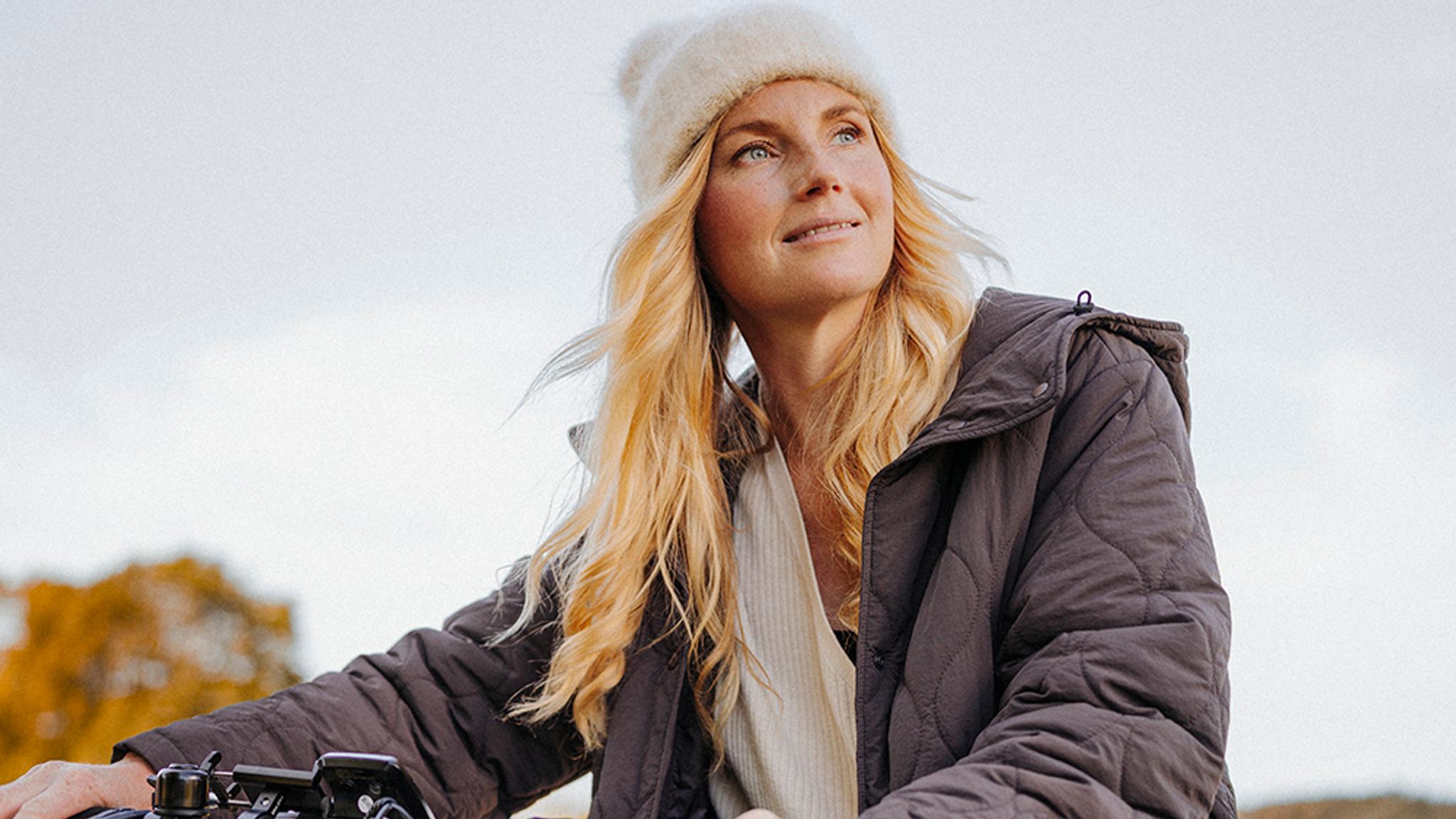 A woman in a cream knit hat and dark quilted jacket smiles and looks up.
