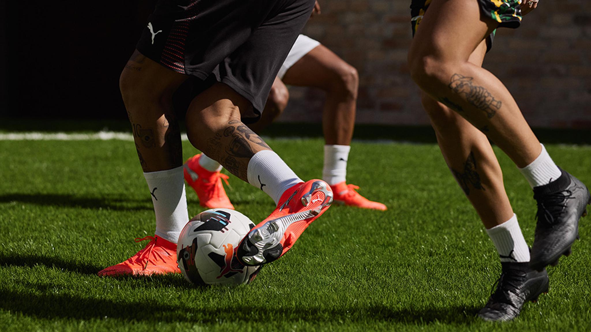 Multiple soccer players' legs on a green field, one foot in an orange cleat dribbling a soccer ball.