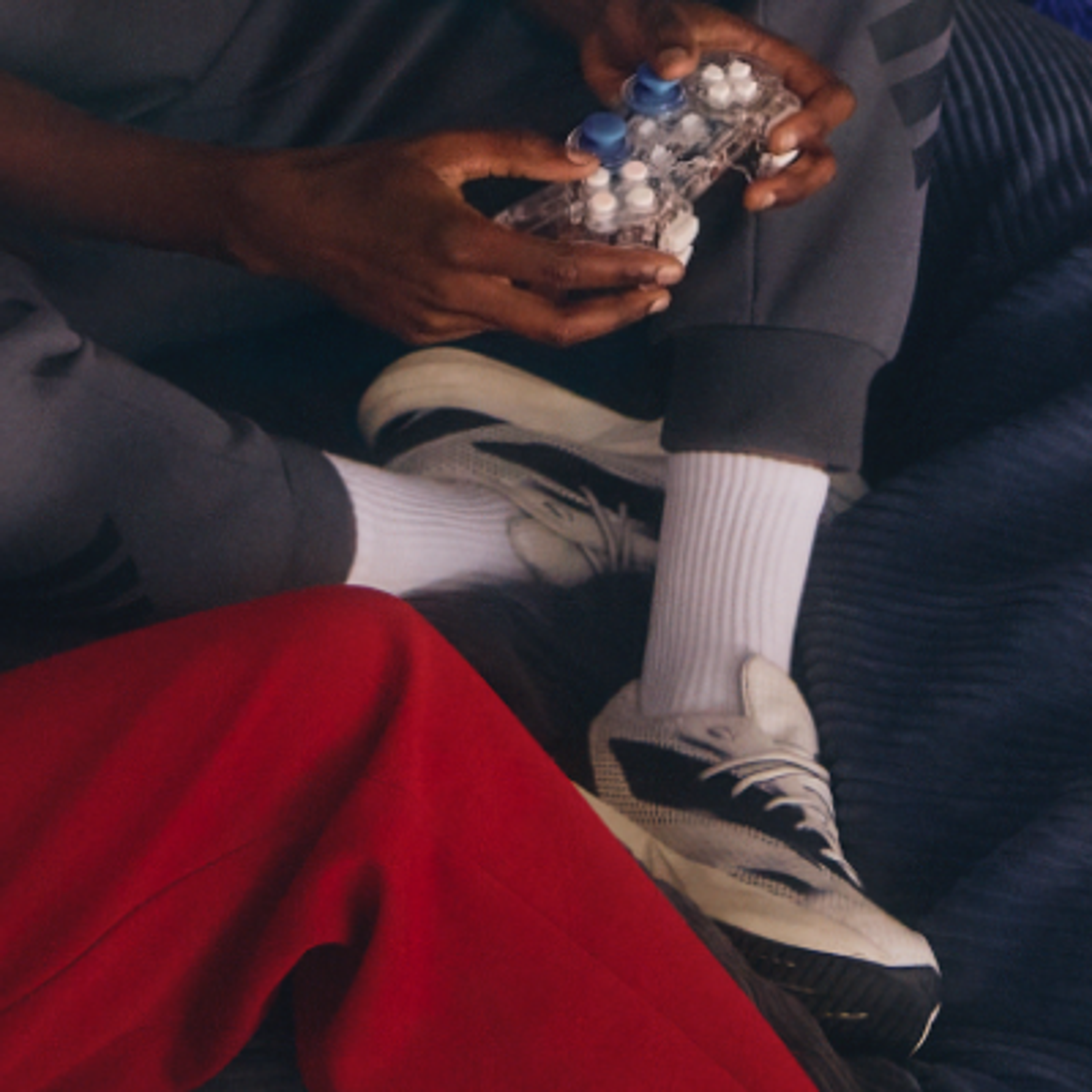 Hands hold a transparent video game controller with blue joysticks and white buttons. The person wears grey sweatpants, white socks, and white sneakers, with a leg in red pants visible in the foreground.