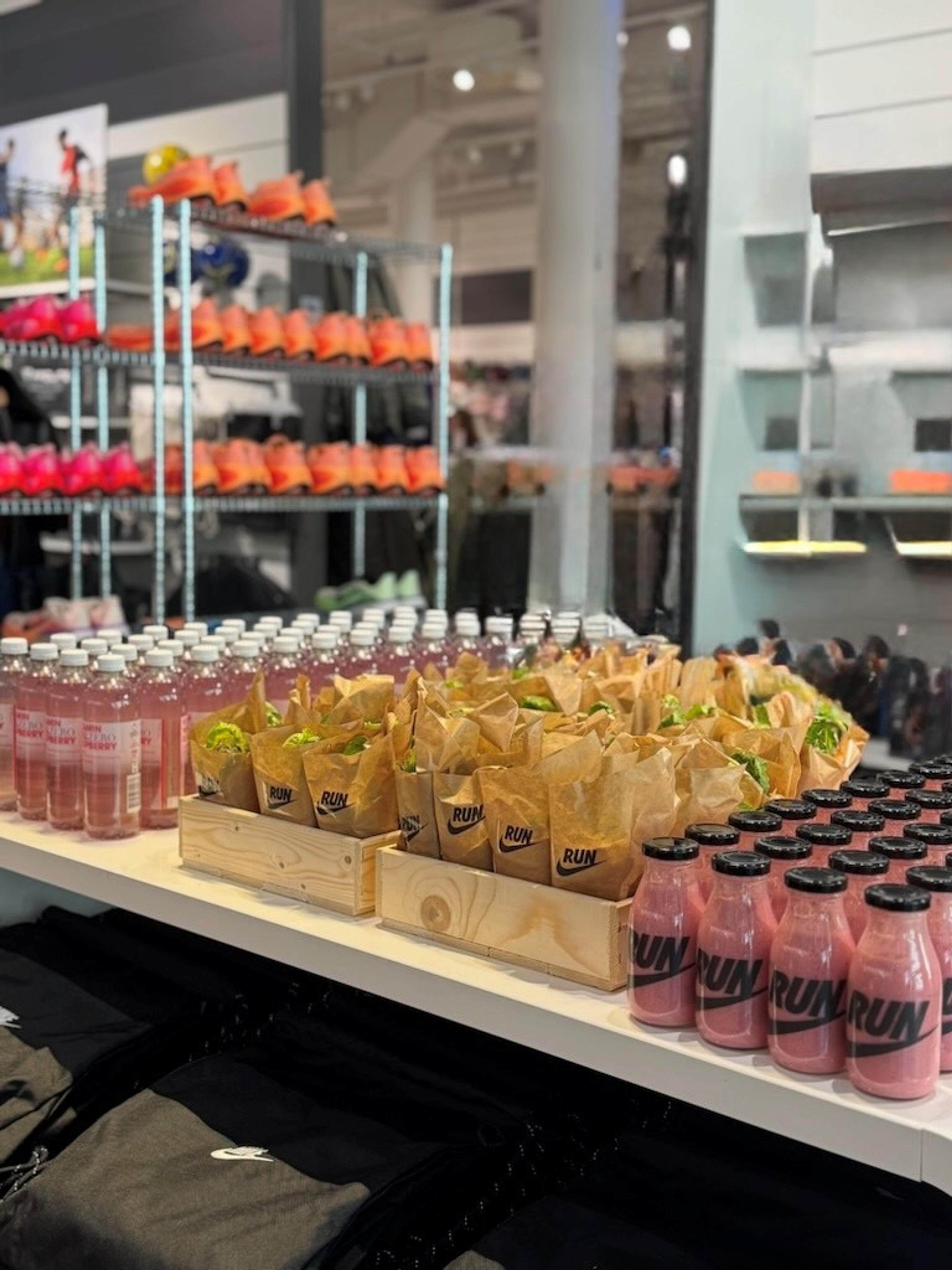 A store display featuring numerous pink "RUN" branded bottled drinks and brown "RUN" paper bags, with shelves of colorful running shoes behind them.