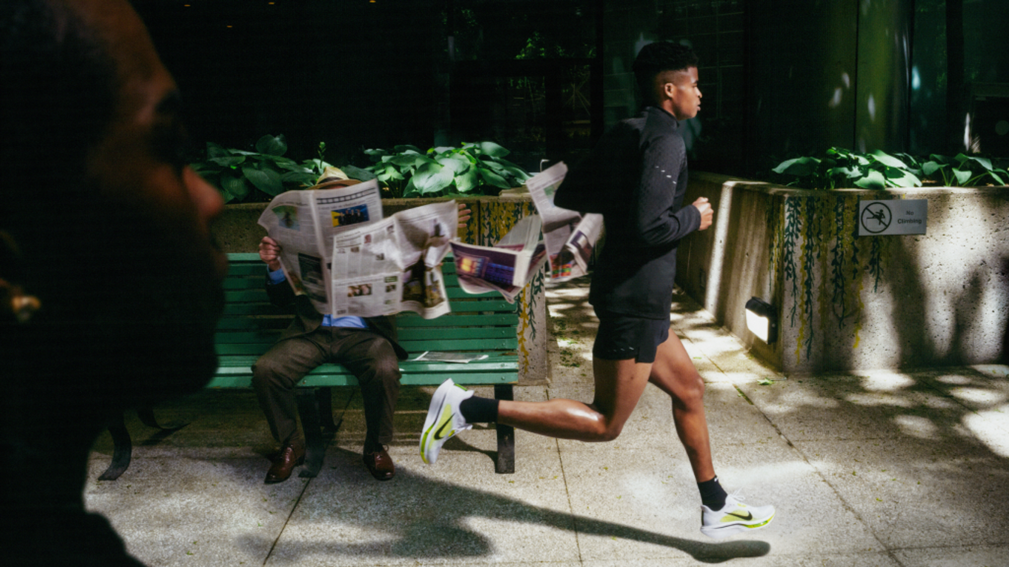A runner in black activewear and bright shoes dashes past a man on a bench reading a fluttering newspaper.
