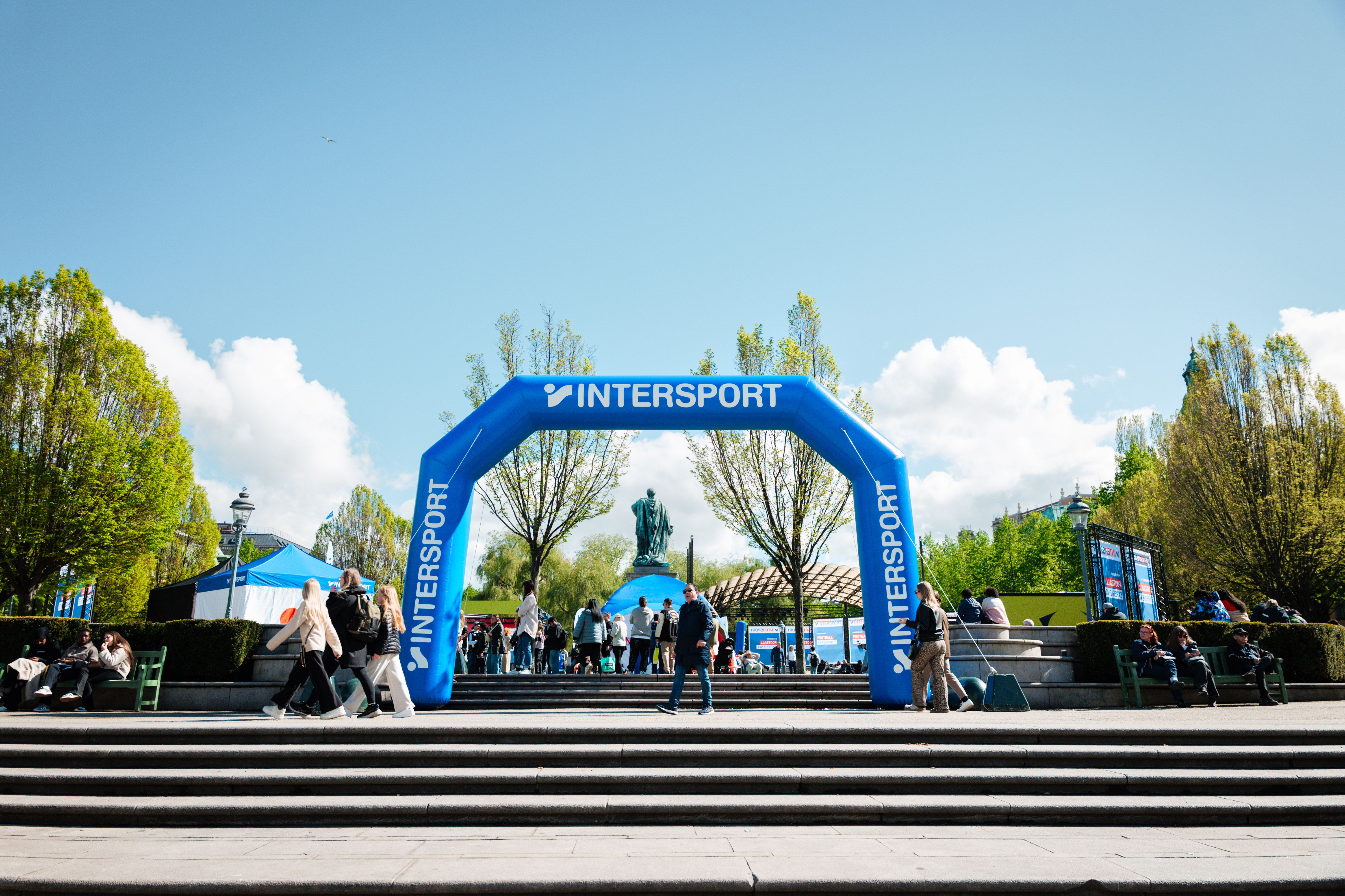 A blue inflatable Intersport archway stands in a bustling public square with people, trees, and a statue in the background.