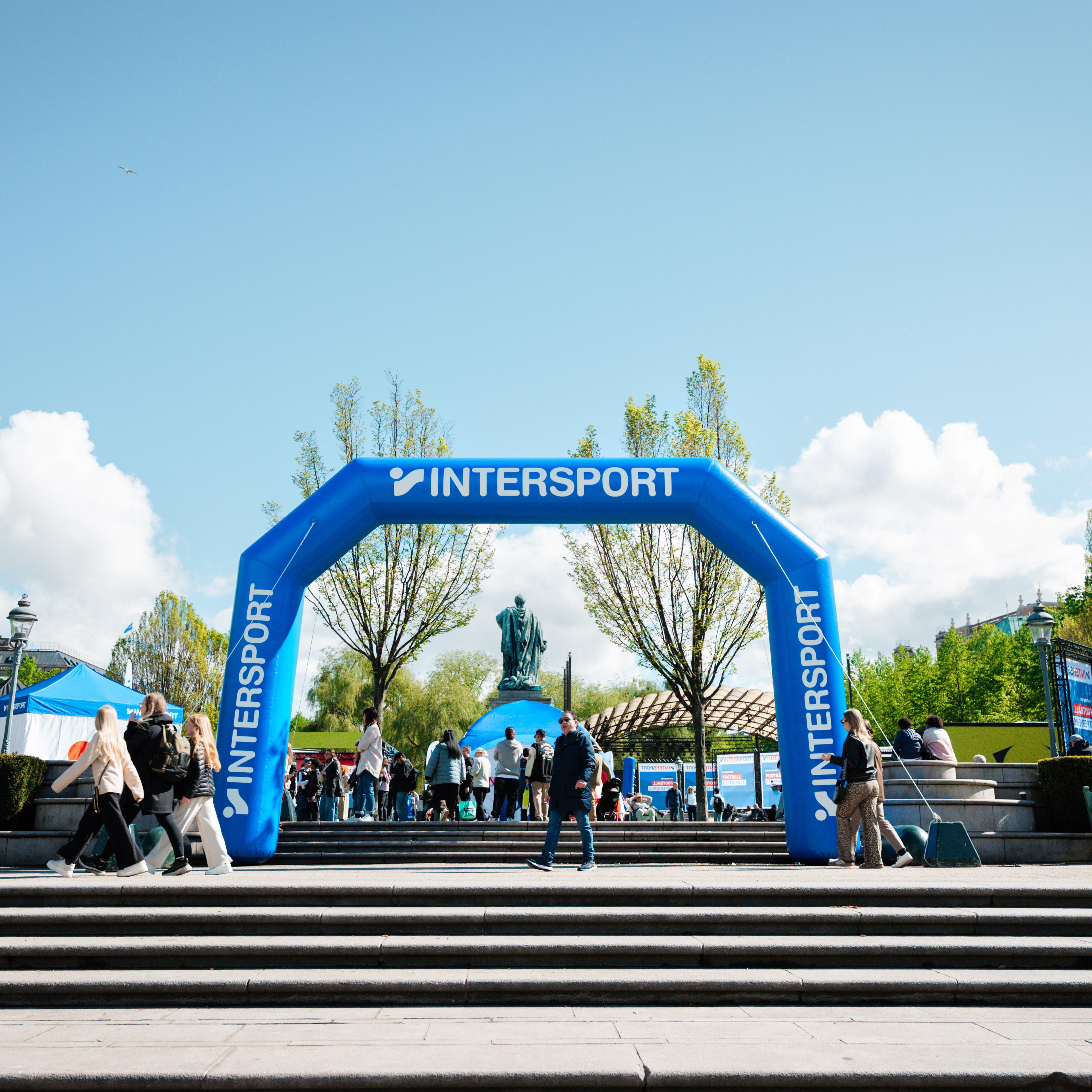 A blue inflatable Intersport archway stands in a bustling public square with people, trees, and a statue in the background.