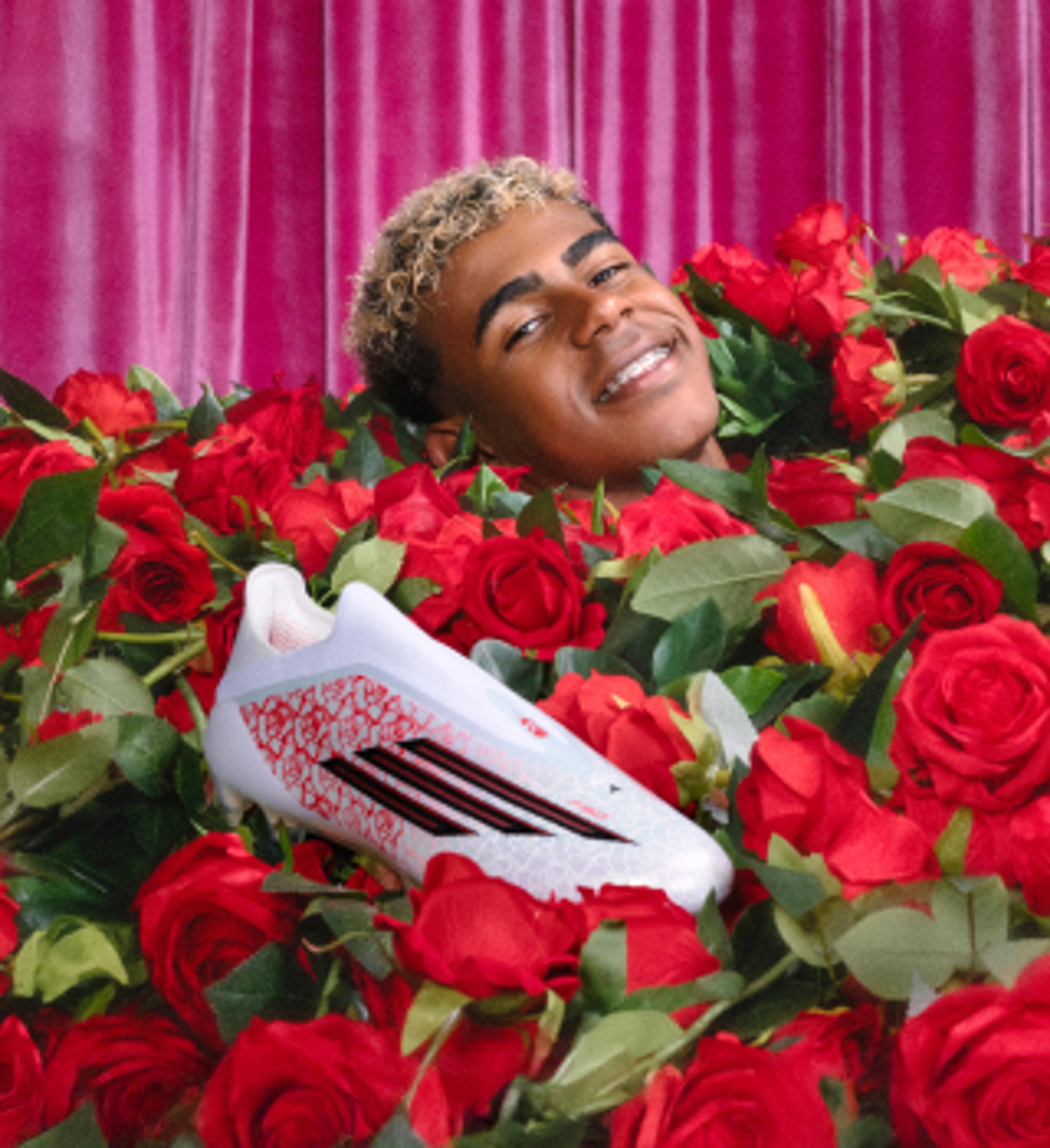 Smiling young man with blonde hair peeking out of red roses next to a white soccer cleat, against a pink curtain.