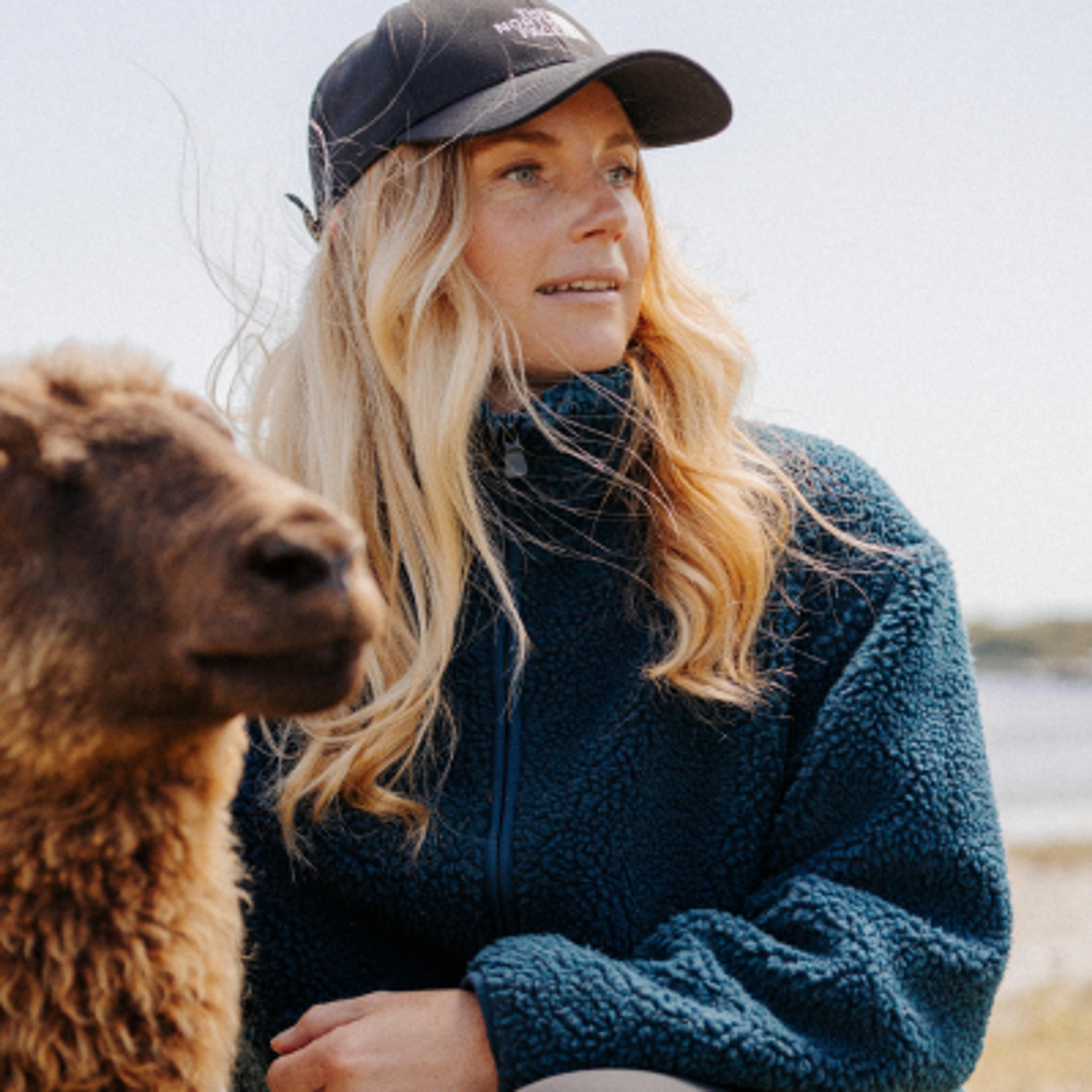 A woman in a black baseball cap and blue fleece jacket looks to the right, with a sheep partially visible beside her on the left.